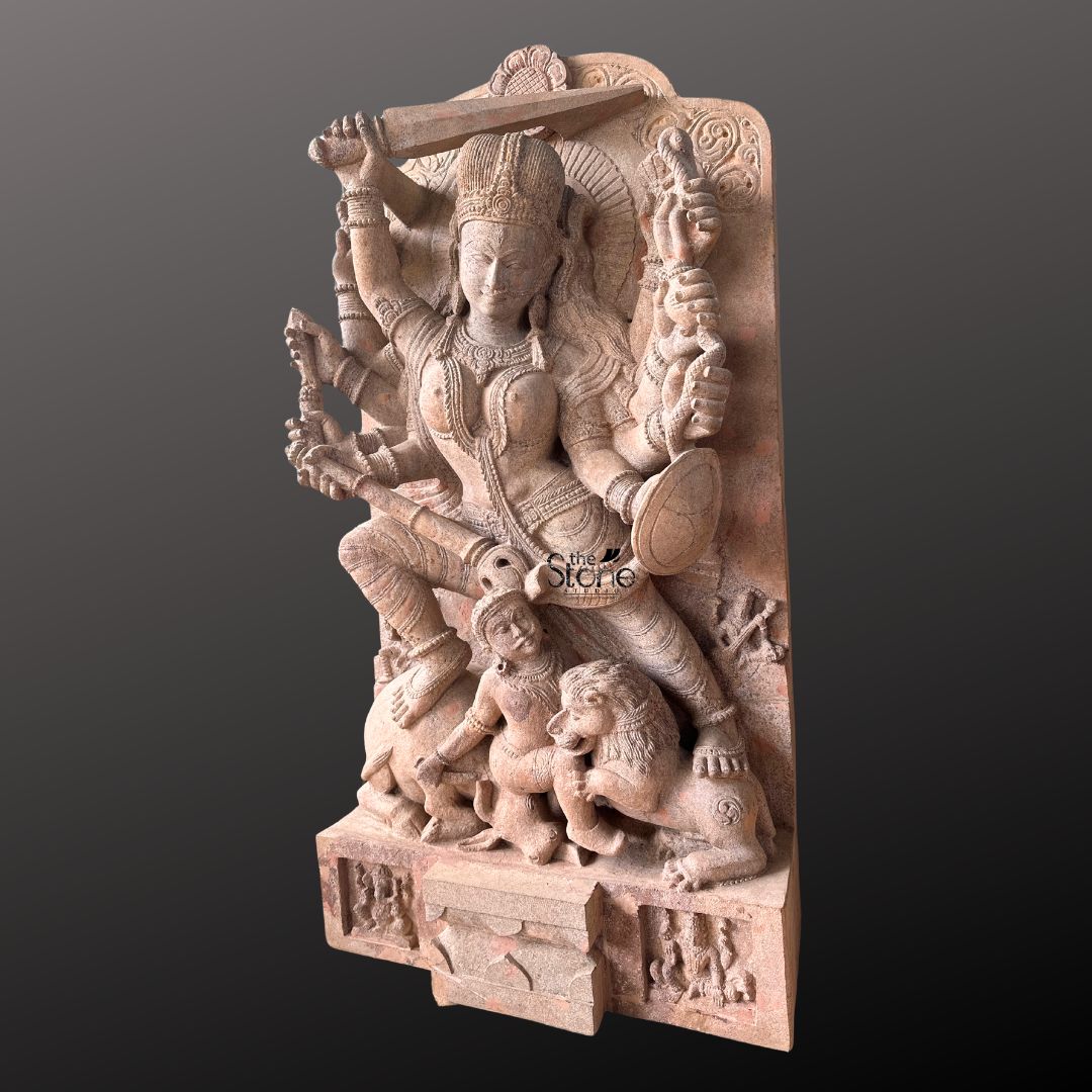 This magnificent stone sculpture carved from a warm-toned pinkish-red sandstone, depicts the Hindu goddess Durga in her powerful form as Mahishasura Mardini the slayer of the buffalo demon. The statue is a high-relief stele showcasing intricate classical Indian artistry. Durga is shown in a dynamic active posture mid-battle with her right foot planted firmly on the severed head of the buffalo demon and her left leg bent. Her primary right hand wields a sword held aloft while her primary left hand holds the tail of the demon who is emerging in human form from the buffalo's carcass. The goddess is depicted with eight arms each holding a symbolic weapon or item including a trident conch disc bow and arrow all rendered in fine detail. She wears elaborate jewelry a detailed crown and a flowing dhoti. The demon Mahishasura is shown in a contorted defeated posture beneath her foot with a lion mount attacking him from the side. The entire sculpture is framed by an ornate archway with floral and geometric carvings and the base includes small relief figures and a central plinth emphasizing the sacred nature of this Mahishasura Mardini Durga statue in Sandstone. The statue is presented against a dark neutral grey background which allows the natural texture and warm color of the sandstone to stand out. The lighting is directional casting soft shadows that enhance the depth and three-dimensionality of the complex carving, showcasing the skilled artisanship. This image is an ideal representation for an article discussing the iconography spiritual significance and cultural heritage of the Maa Durga Mahishasura Mardini Stone Statue.