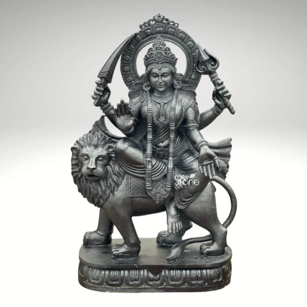 A full-view studio photograph presents a magnificent 4-foot Maa Durga Statue with Lion meticulously hand-carved from dark polished blackstone. The statue depicts the goddess in her divine form seated gracefully upon her powerful roaring lion mount. Maa Durga is portrayed with a serene yet resolute expression adorned with intricate traditional jewelry a detailed crown and flowing robes that reveal the high level of craftsmanship in the blackstone. She is depicted with multiple arms a key feature of her iconography holding various symbolic weapons and attributes. In her raised right hand she holds a khadga and in her raised left a trishula both perfectly rendered in the dark stone. One of her front right hands is raised in the abhaya mudra while one of her front left hands rests in a gesture of blessing. The lion her faithful companion and vahana is sculpted with great strength and detail its mane flowing and mouth open in a powerful roar standing firm on a multi-tiered ornate blackstone pedestal. The entire composition rests upon this decorative base which features carved floral motifs and patterns grounding the monumental figure. The blackstone material exhibits a subtle rich sheen under even, directional studio lighting emphasizing the deep textures and sharp lines of the carvings against a clean neutral light-grey background. The 4-foot height of the statue gives it a commanding and reverential presence, making it a perfect centerpiece for worship or display. This image is an exceptional representation of a Maa Durga Statue with Lion in Black Stone showcasing the traditional artistry and spiritual significance of such a piece. The fine details from the curls of the lion's mane to the delicate patterns on the goddess's garments and the form of her multiple arms, demonstrate the artisan's skill in working with this durable and classic medium. The statue's posture with Durga seated upon the lion symbolizes her divine power and protection conveying a deep sense of reverence and strength suitable for the article's theme. The clean presentation allows the viewer to appreciate the full form and intricate carvings of the 4-foot blackstone murti.