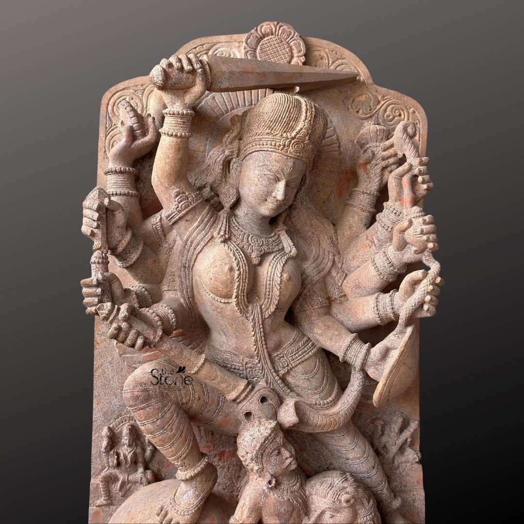 This magnificent stone sculpture carved from a warm-toned sandstone depicts the Hindu goddess Durga in her powerful form as Mahishasura Mardini the slayer of the buffalo demon. The statue is a high-relief stele showcasing intricate classical Indian artistry. Durga is shown in a dynamic active posture, mid-battle with her right foot planted firmly on the severed head of the buffalo demon and her left leg bent. Her primary right hand wields a sword held aloft while her primary left hand holds the tail of the demon who is emerging in human form from the buffalo's carcass. The goddess is depicted with eight arms each holding a symbolic weapon or item including a trident conch disc bow and arrow all rendered in fine detail. She wears elaborate jewelry a detailed crown and a flowing dhoti. The demon Mahishasura is shown in a contorted defeated posture beneath her foot with a lion mount attacking him from the side. The entire sculpture is framed by an ornate archway with floral and geometric carvings and the base includes small relief figures and a central plinth emphasizing the sacred nature of this Mahishasura Mardini Durga statue in Sandstone. The statue is presented against a dark neutral grey background which allows the natural texture and warm color of the sandstone to stand out. The lighting is directional casting soft shadows that enhance the depth and three-dimensionality of the complex carving showcasing the skilled artisanship. This image is an ideal representation for an article discussing the iconography spiritual significance and cultural heritage of the Maa Durga Stone Murti for Temple.