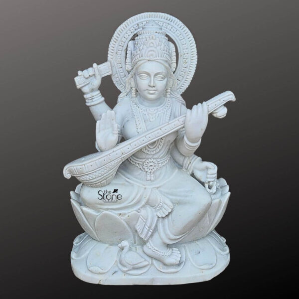 A full-view studio photograph captures a magnificent Maa Saraswati Statue in White Marble standing approximately 24 inches in height. The statue is meticulously hand-carved from a single block of pristine high-quality white Makrana marble giving it a smooth polished finish. The goddess is depicted seated in a graceful Lalitasana pose upon a finely detailed multi-layered lotus throne. Her expression is serene and contemplative with downcast eyes and a gentle smile embodying wisdom and knowledge. She wears elaborate traditional jewelry including necklaces earrings armlets and anklets all intricately carved to look like delicate gold ornaments against the white stone. Maa Saraswati is shown with four arms each holding a symbolic item. In her upper right hand she holds a scripture representing vedic knowledge while her upper left hand holds a mala for spiritual contemplation. Her two primary lower hands gracefully play the veena a stringed musical instrument held diagonally across her body showcasing the artisan's skill in rendering the instrument's details. The folds of her flowing sari are delicately sculpted adding a sense of movement and realism to the marble form. A large ornate halo with intricate geometric patterns frames her head adding to the divine aura of the murti. At the base of the lotus throne her faithful vahana a Hamsa is depicted standing beside her foot. This image is a prime example of traditional Indian stonemasonry and serves as a perfect illustration for an article about acquiring a Maa Saraswati Statue in White Marble. The photograph highlights the exceptional detail and the pure white color of the marble making it easy for a devotee to appreciate the artistry. The clean neutral grey background ensures that the viewer's focus remains entirely on the statue's form texture and traditional iconography. This depiction conveys the spiritual tranquility and cultural significance that such a 24-inch marble murti brings to a home shrine or place of worship.