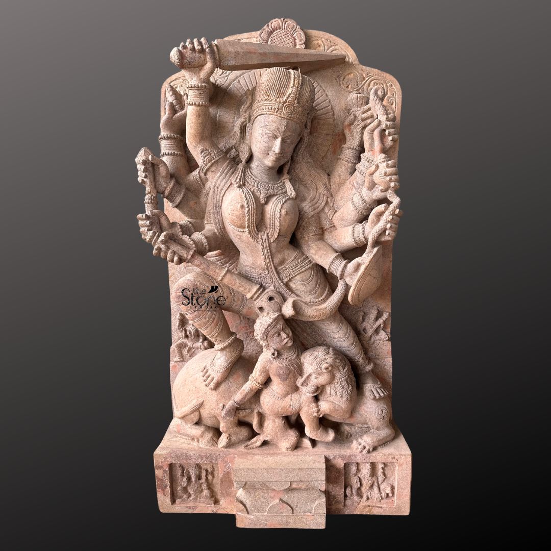 This magnificent stone sculpture carved from a warm-toned pinkish-red sandstone depicts the Hindu goddess Durga in her powerful form as Mahishasura Mardini the slayer of the buffalo demon. The statue is a high-relief stele showcasing intricate classical Indian artistry. Durga is shown in a dynamic, active posture mid-battle with her right foot planted firmly on the severed head of the buffalo demon and her left leg bent. Her primary right hand wields a sword held aloft while her primary left hand holds the tail of the demon who is emerging in human form from the buffalo's carcass. The goddess is depicted with eight arms each holding a symbolic weapon or item including a trident conch disc bow and arrow all rendered in fine detail. She wears elaborate jewelry a detailed crown and a flowing dhoti. The demon Mahishasura is shown in a contorted defeated posture beneath her foot with a lion mount attacking him from the side. The entire sculpture is framed by an ornate archway with floral and geometric carvings and the base includes small relief figures and a central plinth emphasizing the sacred nature of this Mahishasura Mardini Durga statue in Sandstone. The statue is presented against a dark neutral grey background which allows the natural texture and warm color of the sandstone to stand out. The lighting is directional casting soft shadows that enhance the depth and three-dimensionality of the complex carving, showcasing the skilled artisanship. This image is an ideal representation for an article discussing the iconography spiritual significance and cultural heritage of the Mahishasura Mardini Durga Stone Statue.