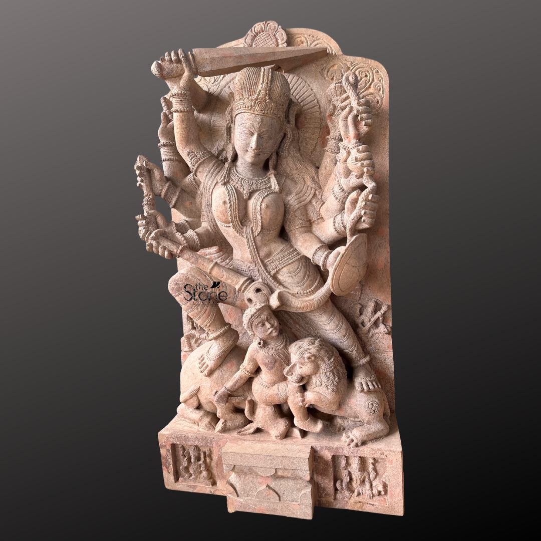 This magnificent stone sculpture carved from a warm-toned pinkish-red sandstone depicts the Hindu goddess Durga in her powerful form as Mahishasura Mardini the slayer of the buffalo demon. The statue is a high-relief stele showcasing intricate classical Indian artistry. Durga is shown in a dynamic active posture mid-battle with her right foot planted firmly on the severed head of the buffalo demon and her left leg bent. Her primary right hand wields a sword held aloft while her primary left hand holds the tail of the demon who is emerging in human form from the buffalo's carcass. The goddess is depicted with eight arms each holding a symbolic weapon or item including a trident conch disc bow and arrow, all rendered in fine detail. She wears elaborate jewelry a detailed crown and a flowing dhoti. The demon Mahishasura is shown in a contorted defeated posture beneath her foot with a lion mount attacking him from the side. The entire sculpture is framed by an ornate archway with floral and geometric carvings and the base includes small relief figures and a central plinth emphasizing the sacred nature of this Mahishasura Mardini Durga statue in Sandstone. The statue is presented against a dark neutral grey background which allows the natural texture and warm color of the sandstone to stand out. The lighting is directional casting soft shadows that enhance the depth and three-dimensionality of the complex carving, showcasing the skilled artisanship. This image is an ideal representation for an article discussing the iconography spiritual significance and cultural heritage of the Mahishasura Mardini Statue in Stone.