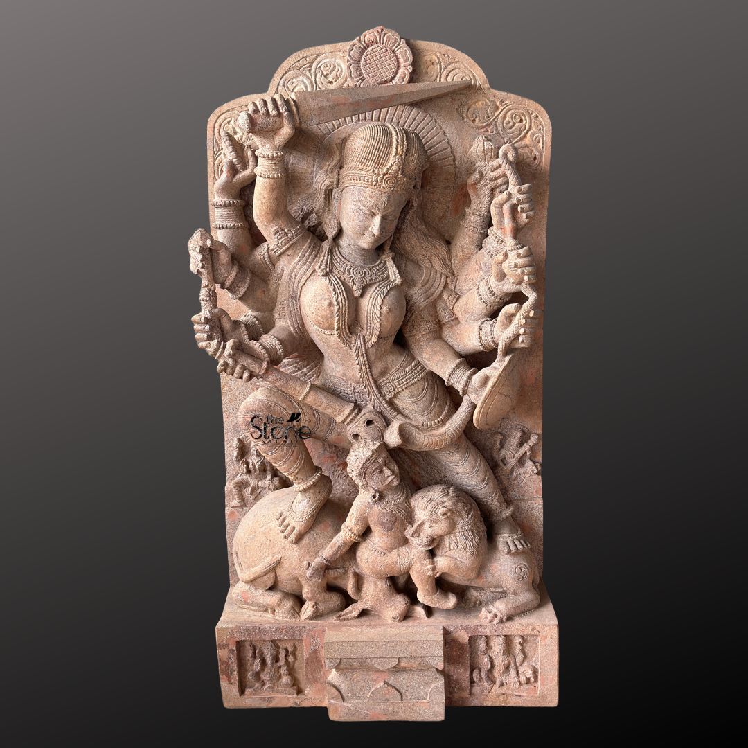This magnificent stone sculpture carved from warm-toned pinkish-red sandstone depicts the Hindu goddess Durga in her powerful form as Mahishasura Mardini the slayer of the buffalo demon. The statue is a high-relief stele showcasing intricate classical Indian artistry. Durga is shown in a dynamic active posture mid-battle with her right foot planted firmly on the severed head of the buffalo demon and her left leg bent. Her primary right hand wields a sword held aloft while her primary left hand holds the tail of the demon who is emerging in human form from the buffalo's carcass. The goddess is depicted with eight arms each holding a symbolic weapon or item including a trident conch disc bow and arrow all rendered in fine detail. She wears elaborate jewelry a detailed crown and a flowing dhoti. The demon Mahishasura is shown in a contorted defeated posture beneath her foot with a lion mount attacking him from the side. The entire sculpture is framed by an ornate archway with floral and geometric carvings and the base includes small relief figures and a central plinth emphasizing the sacred nature of this Mahishasura Mardini Durga statue in Sandstone. The statue is presented against a dark neutral grey background which allows the natural texture and warm color of the sandstone to stand out. The lighting is directional casting soft shadows that enhance the depth and three-dimensionality of the complex carving showcasing the skilled artisanship. This image is an ideal representation for an article discussing the iconography spiritual significance and cultural heritage of the Mahishasura Mardini Stone Murti for Temple.