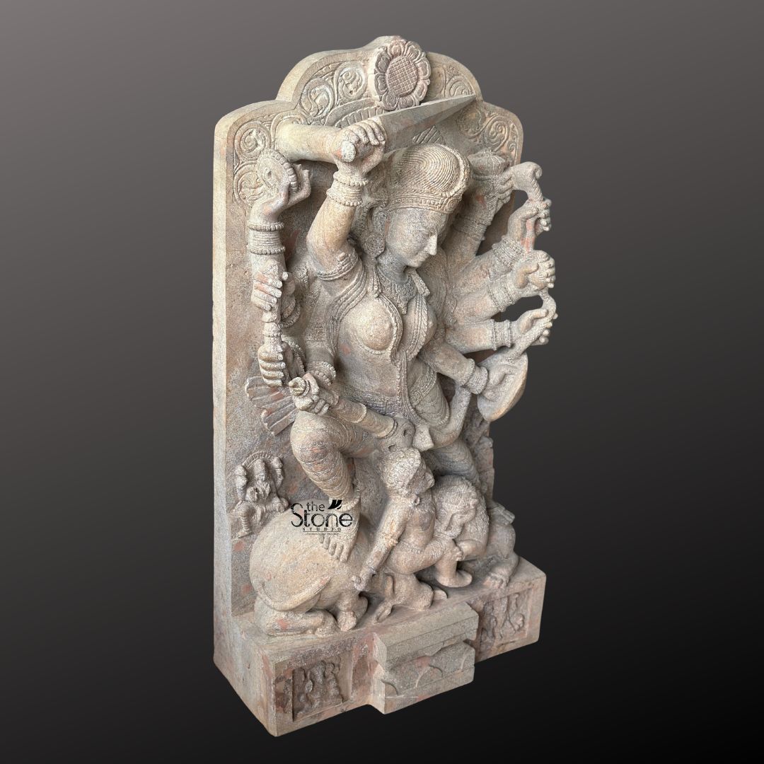 This magnificent stone sculpture carved from a warm-toned pinkish-red sandstone, depicts the Hindu goddess Durga in her powerful form as Mahishasura Mardini the slayer of the buffalo demon. The statue is a high-relief stele showcasing intricate classical Indian artistry. Durga is shown in a dynamic active posture mid-battle with her right foot planted firmly on the severed head of the buffalo demon and her left leg bent. Her primary right hand wields a sword held aloft while her primary left hand holds the tail of the demon who is emerging in human form from the buffalo's carcass. The goddess is depicted with eight arm each holding a symbolic weapon or item including a trident conch disc bow and arrow all rendered in fine detail. She wears elaborate jewelry a detailed crown and a flowing dhoti. The demon Mahishasura is shown in a contorted defeated posture beneath her foot with a lion mount attacking him from the side. The entire sculpture is framed by an ornate archway with floral and geometric carvings, and the base includes small relief figures and a central plinth emphasizing the sacred nature of this Mahishasura Mardini Durga statue in Sandstone. The statue is presented against a dark neutral grey background which allows the natural texture and warm color of the sandstone to stand out. The lighting is directional casting soft shadows that enhance the depth and three-dimensionality of the complex carving, showcasing the skilled artisanship. This image is an ideal representation for an article discussing the iconography spiritual significance and cultural heritage of the Mahishasuramardini Durga Devi Stone Idol.