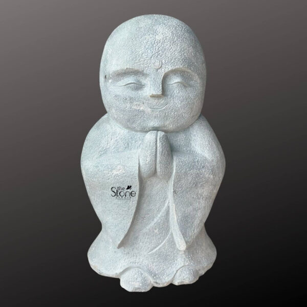 This is a full-view photograph of a charming hand-carved stone statue of a Buddhist monk standing 3 feet tall and designed as a serene focal point for garden decor. The statue is crafted from a high-quality speckled blackstone which gives it a dark mottled grey-blue appearance with a slightly rough tactile texture. The monk is depicted in a seated or kneeling posture with his body slightly rounded and his feet tucked beneath him. His hands are brought together at the chest in the traditional Anjali Mudra or Namaste gesture of greeting and reverence perfectly capturing the essence of the article's subject. The monk's face is rendered in a simple stylized and endearing manner with eyes gently closed in a peaceful meditative smile and subtly defined features. He has a smooth shaven head and the overall impression is one of calm devotion. The carving extends to the details of his robes which are represented with clean flowing lines carved into the stone showing deep folds and sleeves that drape over his hands and lap conveying a sense of weight and texture inherent to the blackstone material. The statue is presented against a seamless dark grey studio background that fades to black isolating the figure and emphasizing its form and texture. The lighting is soft and even illuminating the statue from the front and slightly to the side, which casts gentle shadows that define its three-dimensional quality and highlight the rough porous nature of the blackstone. The base of the statue is wide and stable making it suitable for placement directly on the ground a garden pedestal or a tiered altar.