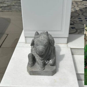 This is a detailed eye-level photograph of a traditional hand-carved Nandi statue crafted from a dark textured blackstone. The statue which measures 18 inches in length depicts Nandi the sacred bull and vehicle of Lord Shiva, in a reverent, seated posture. Nandi is shown facing forward with folded legs and a calm serene expression. The sculpture is richly adorned with intricate carvings including decorative bells harnesses and garlands that cover its head neck and body showcasing the artisan's skill in creating fine details in the hard stone. The blackstone material has a matte slightly porous finish that enhances the texture of the carvings. The Nandi statue is positioned as a decorative focal point in a modern upscale outdoor or semi-outdoor setting likely a home entrance or garden patio in Chandigarh. It rests securely on a clean polished white marble surface that serves as a pedestal creating a striking contrast between the dark stone of the sculpture and the light reflective marble. The statue is oriented towards the viewer presenting a complete view of its form and ornamentation. The background consists of elements common to a contemporary Chandigarh residence. Behind the statue is a large smooth white panel possibly a modern architectural wall or pillar. To the left a paved area leads towards a wall clad in stacked grey slate stone adding texture and depth to the background. To the right a small glimpse of a lush green garden with tropical plants provides a soft organic contrast to the hard lines of the architecture and the stone sculpture. Bright natural daylight illuminates the scene from the right casting defined shadows that emphasize the three-dimensional quality and deep reliefs of the Nandi carving.