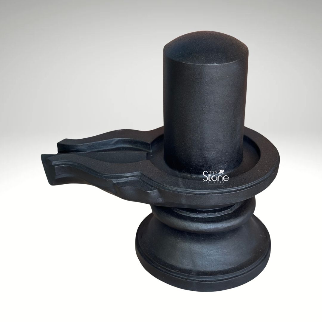 A three-quarter studio photograph captures a traditional Blackstone Shivling statue meticulously hand-carved from a single piece of dark rich natural blackstone. The entire murti rests on a simple rounded pedestal base, providing a stable foundation. Rising from this base is the yoni pedestal featuring a perfectly centered smooth shivling shaft. The dark polished surface of the stone absorbs and reflects light subtly emphasizing the heavy solid presence of the murti and the tactile quality of the material. The clean neutral background ensures that the viewer's attention is entirely on the sculpture's form texture and traditional iconography. The artistry of this Natural Black Stone Shiva Lingam is evident in the fine detail and traditional craftsmanship. The smooth cylindrical linga represents the formless aspect of Lord Shiva while the yoni base symbolizes the creative energy of Goddess Parvati. The harmonious union of these two elements is central to Hindu philosophy. To the side the intricately carved pranala extends outwards, showing precise channels and traditional decorative motifs that are characteristic of fine Indian stonemasonry designed to catch and channel ritual offerings of water milk and other liquids. This murti serves as an exemplary representation of a Natural Black Stone Shiva Lingam highlighting the specific craftsmanship and quality of the blackstone used. The photograph effectively communicates the cultural significance spiritual symbolism and physical beauty of the devotional object making it a valuable visual reference for devotees and collectors interested in acquiring a traditionally crafted Shivling for a home or temple shrine. The high-quality lighting enhances the deep texture of the stone showcasing the skill of the artisan in every curve and line.