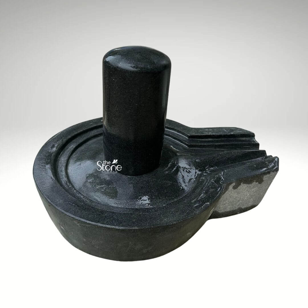 A centered eye-level studio photograph presents a meticulously hand-carved Natural Black Stone Shivling complete with its Yoni base set against a clean light grey gradient background. The entire sacred artifact is carved from a single block of dense dark black granite showcasing the stone's natural finely crystalline texture and deep rich color. The central element is the cylindrical Shivling representing the formless aspect of Lord Shiva which rises smoothly and vertically from the center of the circular Yoni pedestal. The top of the Shivling is gently domed and its surface is highly polished reflecting ambient light and creating a subtle sheen that contrasts with the matte finish of the surrounding elements. The Yoni base symbolizing the feminine creative energy of Goddess Parvati is a circular basin-like structure surrounding the base of the Shivling. It features a clearly defined raised circular rim and a well-proportioned projecting spout to the right side designed to channel ceremonial offerings of water or milk away from the deity. The entire Yoni base is also meticulously carved from the same dark black stone with precise concentric rings enhancing its form and balance. The carving exhibits traditional craftsmanship with smooth flowing curves and precise edges that give the Shivling and Yoni a harmonious and integrated appearance conveying a sense of ancient sanctity. This image serves as a prime visual reference for an article discussing the traditional iconography spiritual significance and selection of a Natural Black Stone Shivling for home temples. The detailed depiction of the dark granite the specific shape of the Yoni and the presence of the spout are all key elements relevant to the article's content. The single-piece construction from durable black granite makes it a sought-after item for daily worship and ritualistic abhishekam. The high-quality finish and accurate proportions make this particular statue an excellent representation of a traditional Hindu devotional murti suitable for creating a sacred space.