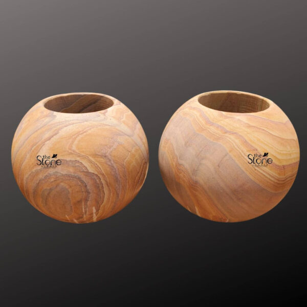 A studio photograph presents a pair of identical spherical sandstone planter pots arranged side-by-side against a softly graduated dark grey background. Each pot is hand-carved from a single block of natural sandstone showcasing a rich warm honey-gold base color interwoven with distinctive flowing layered strata patterns in shades of cream beige and light brown. This unique natural veining gives each piece a one-of-a-kind artisanal appearance. The pots are perfectly spherical with a smooth tactile matte finish that emphasizes the inherent beauty of the stone. The top of each sphere is hollowed out to form a perfectly circular deep basin approximately half the diameter of the pot designed to hold a plant and soil. The rim of the opening is clean and smooth leading down into the planting chamber. The texture of the sandstone is clearly visible exhibiting a fine-grained slightly porous quality that is both durable and aesthetically pleasing. The substantial weight and solid form of the pots convey a sense of stability and quality craftsmanship. These natural sandstone round planter pots are specifically designed to be a sophisticated and organic addition to any home decor or garden setting. The earthy colors and flowing patterns of the stone complement a wide range of plant life from succulents and cacti to small flowering plants or ferns. The spherical shape is a timeless and elegant design choice that brings a soft natural element to modern or traditional spaces. The durability of the sandstone makes them suitable for both indoor and outdoor use ensuring they remain a beautiful and functional piece for years to come. Their artisanal quality and natural material highlight a commitment to sustainable and elegant living.