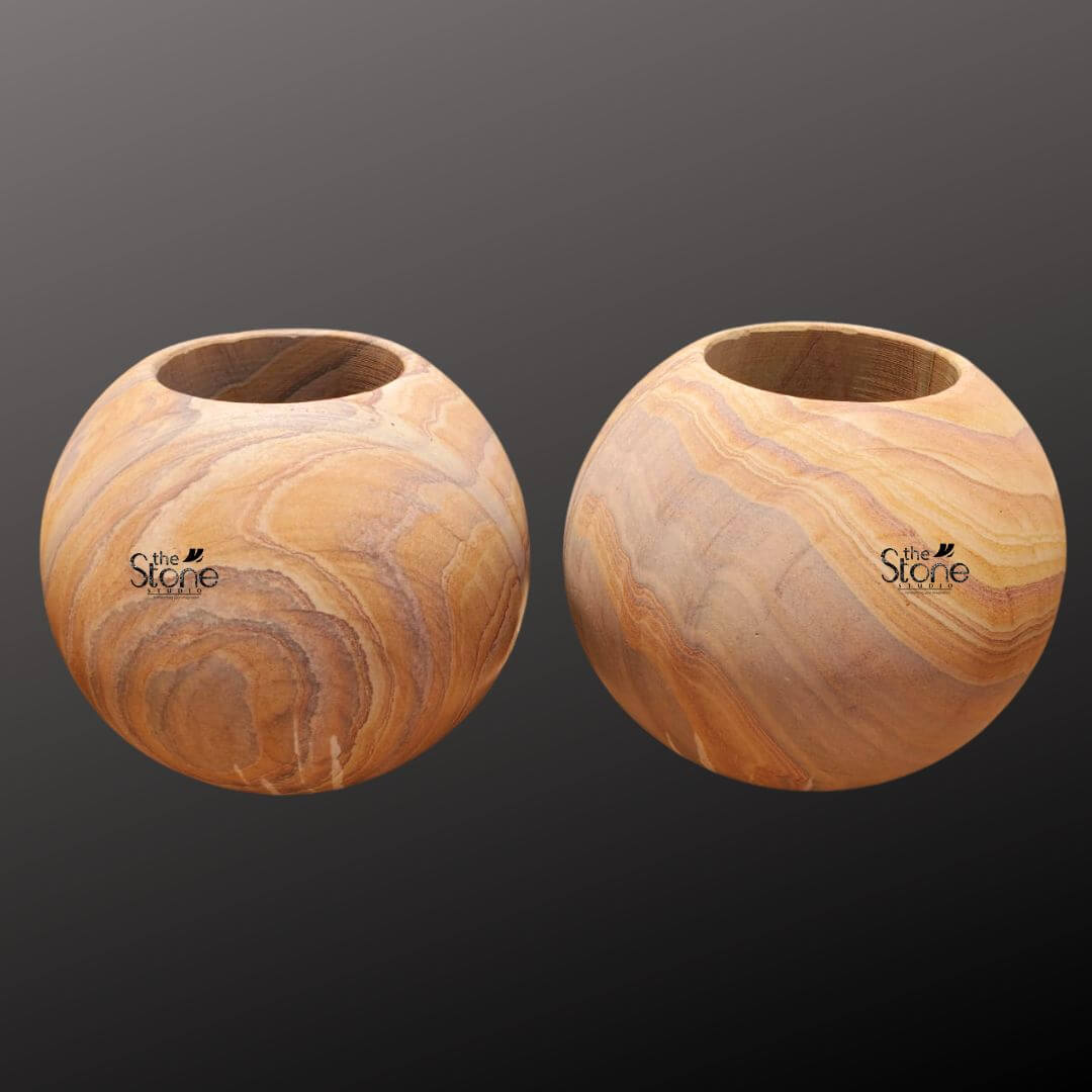 A studio photograph presents a pair of identical spherical sandstone planter pots arranged side-by-side against a softly graduated dark grey background. Each pot is hand-carved from a single block of natural sandstone showcasing a rich warm honey-gold base color interwoven with distinctive flowing layered strata patterns in shades of cream beige and light brown. This unique natural veining gives each piece a one-of-a-kind artisanal appearance. The pots are perfectly spherical with a smooth tactile matte finish that emphasizes the inherent beauty of the stone. The top of each sphere is hollowed out to form a perfectly circular deep basin approximately half the diameter of the pot designed to hold a plant and soil. The rim of the opening is clean and smooth leading down into the planting chamber. The texture of the sandstone is clearly visible exhibiting a fine-grained slightly porous quality that is both durable and aesthetically pleasing. The substantial weight and solid form of the pots convey a sense of stability and quality craftsmanship. These natural sandstone round planter pots are specifically designed to be a sophisticated and organic addition to any home decor or garden setting. The earthy colors and flowing patterns of the stone complement a wide range of plant life from succulents and cacti to small flowering plants or ferns. The spherical shape is a timeless and elegant design choice that brings a soft natural element to modern or traditional spaces. The durability of the sandstone makes them suitable for both indoor and outdoor use ensuring they remain a beautiful and functional piece for years to come. Their artisanal quality and natural material highlight a commitment to sustainable and elegant living.