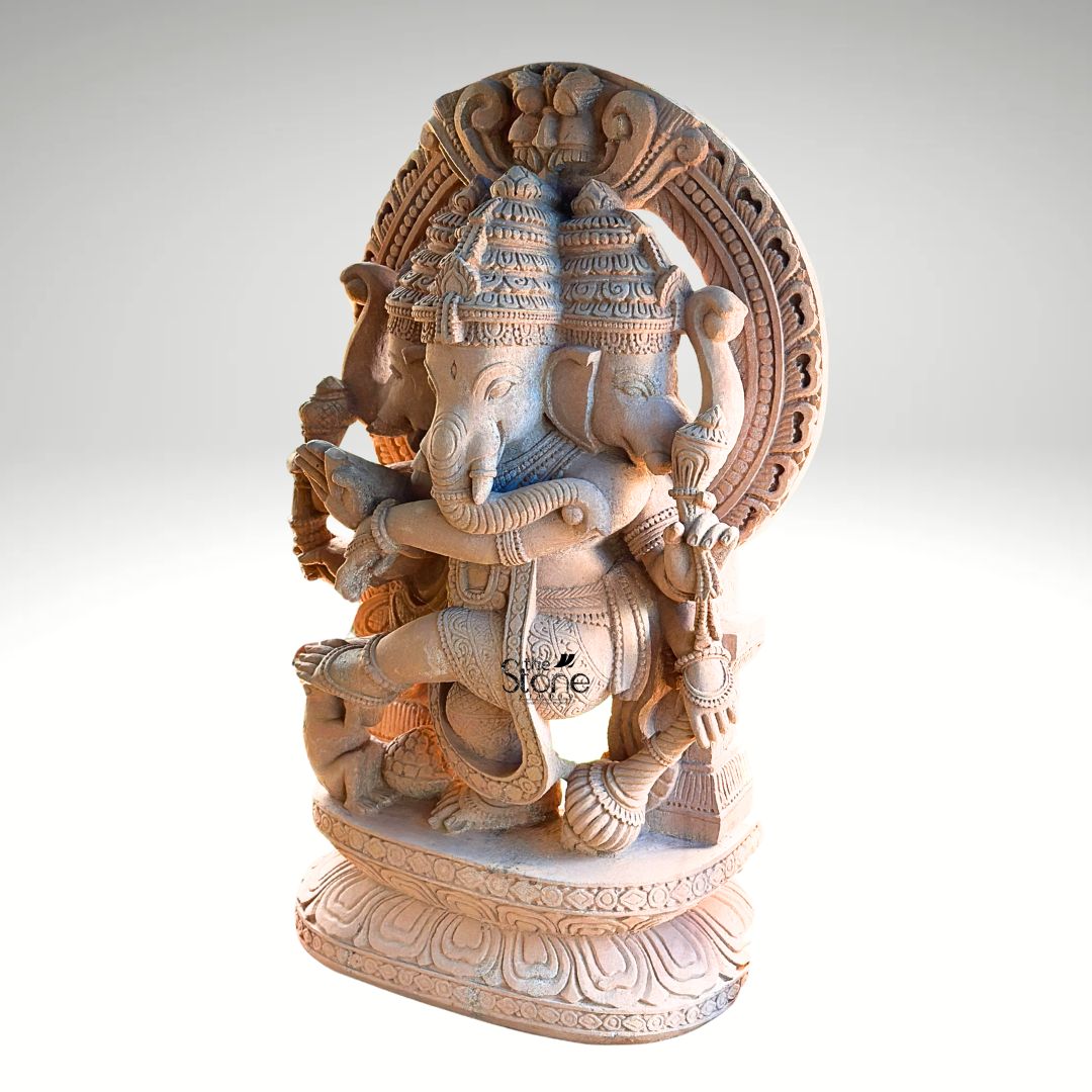 A full-view studio photograph captures a magnificent 4-foot-tall Nritya Ganapathi Idol hand-carved from warm earthy sandstone. The statue is depicted in a dynamic dancing pose with Ganesha's left leg raised and bent gracefully across his body conveying a sense of divine rhythm and movement. He is adorned with elaborate traditional jewelry including multiple necklaces armlets and a sacred thread all intricately sculpted from the same durable stone. The natural porous texture and warm reddish-brown color of the sandstone are beautifully highlighted, giving the sculpture a rich tactile quality. This unique murti features three heads with Ganesha's primary elephant face looking forward flanked by two identical elephant heads facing left and right all wearing ornate crowns. Ganesha is shown with eight arms each holding a symbolic attribute such as an ankusha a pasha and a modak with his trunk elegantly curved to taste a sweet from his lower left hand. The sculpture rests on a finely detailed multi-layered lotus throne grounding the monumental figure. At Ganesha's feet a small figure of his vahana the mouse is depicted looking upwards in reverence. The entire form of the Nritya Ganapathi is framed by a spectacular large ornate Prabhavali arch that rises behind him. The arch is richly carved with traditional patterns including stylized kirtimukha motifs at the apex and floral designs along its length. This image serves as an exemplary representation of traditional Indian stonemasonry and is a perfect illustration for an article discussing the significance of a Nritya Ganapathi Idol with Prabhavali Arch. The photograph highlights the exceptional detail the imposing scale, and the warm natural color of the sandstone making it easy for a devotee or collector to appreciate its spiritual energy and artistic grace.