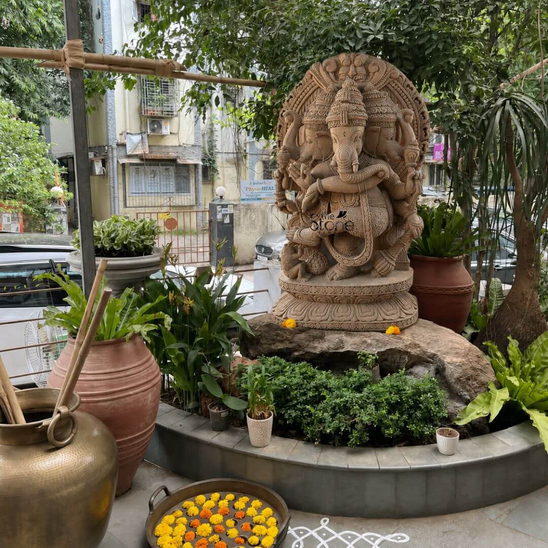 A candid daylight photograph captures a magnificent 4-foot-tall Nritya Ganapathi Three Face Stone Sculpture, hand-carved from a single block of warm earthy sandstone. The statue is the majestic centerpiece of an outdoor garden shrine in an urban setting placed upon a rough-hewn rock base and surrounded by an abundance of lush green potted plants including large palms ferns and shrubs. The sculpture depicts Lord Ganesha in a dynamic dancing pose with his left leg raised and bent gracefully across his body conveying a sense of divine rhythm and movement. This unique murti features three heads with Ganesha's primary elephant face looking forward flanked by two identical elephant faces facing left and right all wearing ornate crowns. Ganesha is shown with eight arms each holding a symbolic attribute such as an ankusha a pasha and a modak with his trunk elegantly curved to taste a sweet from his lower left hand. The sculpture rests on a finely detailed multi-layered lotus throne, grounding the monumental figure. Behind him a large ornate halo with traditional patterns frames his head adding to the divine aura. This image serves as an exemplary representation of traditional Indian stonemasonry and is a perfect illustration for an article discussing the significance of a Nritya Ganapathi Three Face Stone Sculpture. The photograph highlights the exceptional detail and the imposing presence of the 4-foot sandstone statue within its naturalized urban environment making it easy for a devotee or collector to appreciate its spiritual energy and artistic grace. The setting is complemented by a large brass water pot a floating bowl of yellow and orange marigold flowers and traditional rangoli patterns on the stone-paved ground conveying the significant role such a murti plays as a focal point for devotion and cultural heritage.