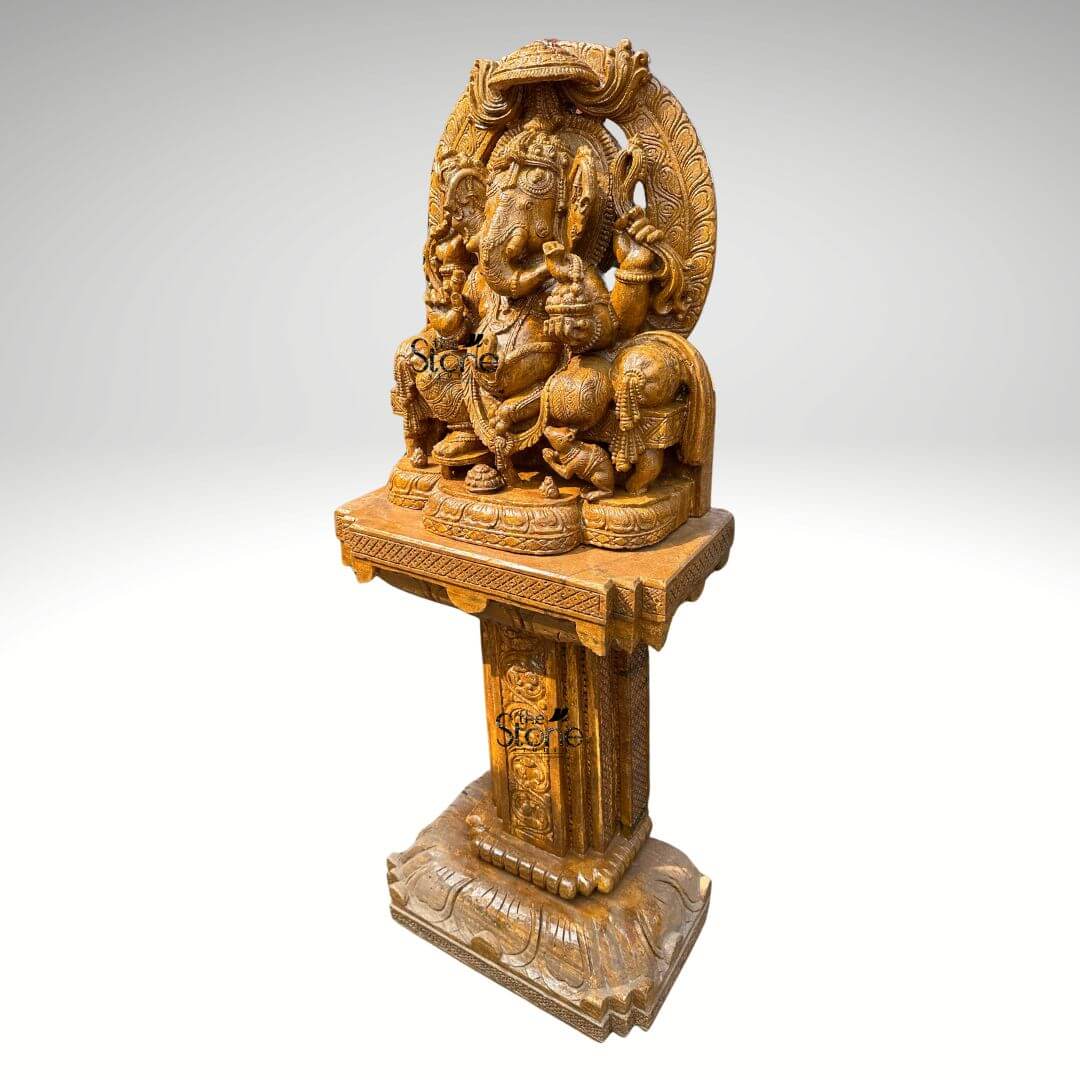 This is a full-view photograph of a magnificent hand-carved brown sandstone statue of Lord Ganesha elevated and displayed upon a matching ornately carved sandstone pedestal designed as a grand focal point for outdoor garden decor. The statue depicts Ganesha in a traditional seated posture known as Vighneshvara upon a richly carved throne. He is adorned with intricate traditional jewelry a detailed crown and garlands all carved directly from the warm textured brown sandstone. Ganesha has four arms holding symbolic attributes including an axe and a goad with his primary right hand in the Abhaya mudra of blessing. His form is robust and detailed with a serene facial expression and deeply carved reliefs that showcase artisanal skill. The Ganesha statue rests upon a substantial square sandstone pedestal base which is richly carved on all visible sides with traditional Indian motifs including decorative floral patterns miniature figural reliefs and architectural elements that echo the artistry of the main sculpture. The pedestal provides a stable and elegant foundation raising the deity to a prominent height suitable for worship or prominent display as an outdoor decor piece. The entire ensemble—statue throne and pedestal—is crafted from the same consistent brown sandstone featuring a beautiful uniform natural grain and a finely textured matte finish that emphasizes the depth and complexity of the hand-carvings making it durable for outdoor environments. The statue and pedestal are presented against a clean seamless light-grey studio background that fades to white isolating the object and allowing its form color and intricate details to be the sole focus. The lighting is soft even and directional illuminating the scene from the front and slightly to the side to cast gentle shadows that define the three-dimensional quality and deep reliefs of the sandstone carving. The overall composition conveys a sense of spiritual reverence traditional artistry and substantial craftsmanship inherent to this type of large Indian stone sculpture.
