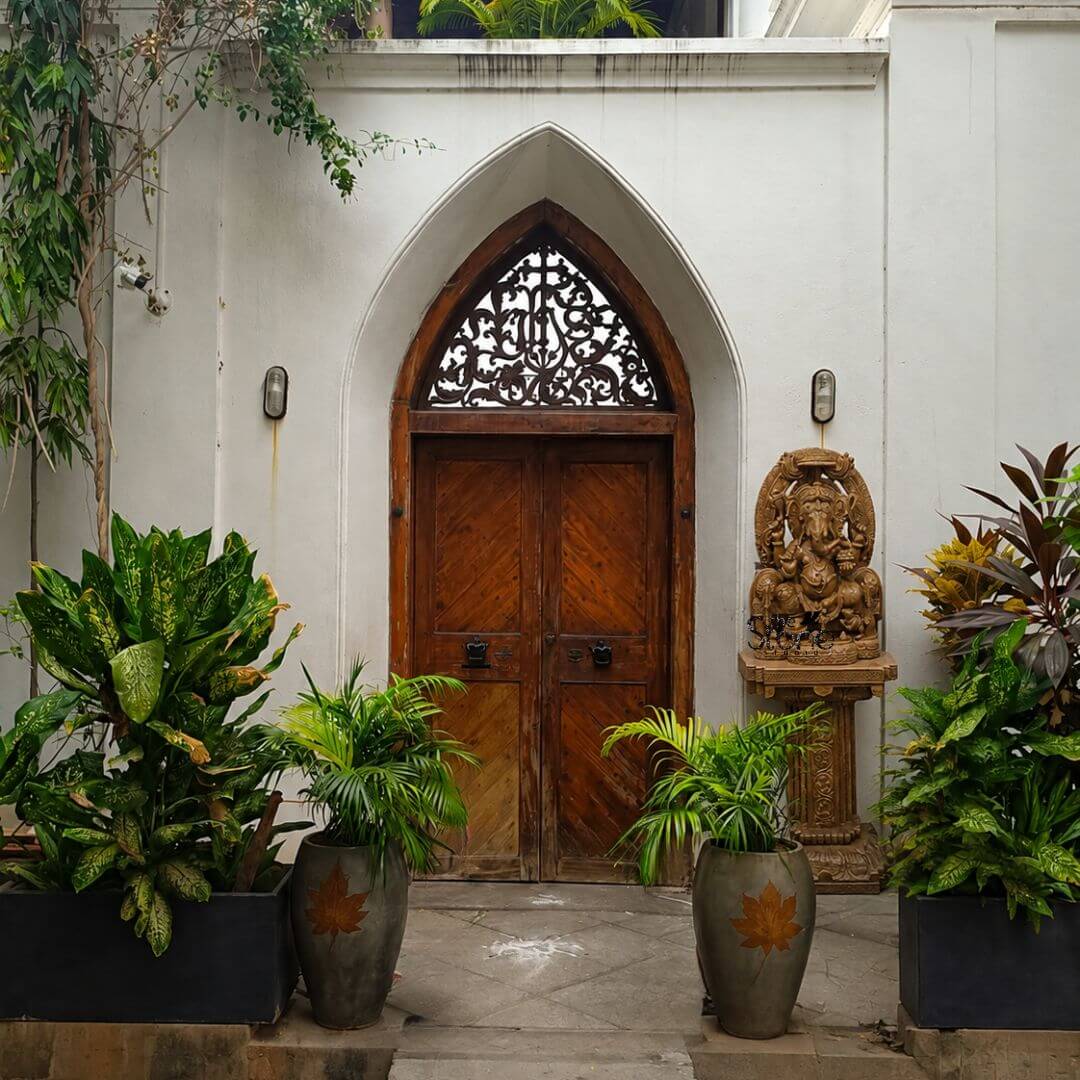 This is a centered eye-level photograph of a magnificent, hand-carved brown sandstone statue of Lord Ganesha positioned as an elegant centerpiece at a sophisticated villa entrance. The statue, standing approximately four feet tall is crafted from a warm textured brown sandstone and depicts Ganesha in a traditional seated posture known as Vighneshvara upon a richly carved multi-tiered throne. He is adorned with intricate traditional jewelry a detailed crown and garlands all carved directly from the stone with deep reliefs. Ganesha has four arms, holding symbolic attributes including an axe and a goad with his primary right hand in the Abhaya mudra of blessing. The Ganesha statue rests upon an ornate carved sandstone pedestal base which is richly decorated with traditional Indian motifs echoing the art style of the main sculpture. This entire assembly is situated on a paved stone courtyard flanked symmetrically by large well-manicured tropical plants in varied planters. On the left a large trough-style planter holds lush croton and palm plants while a smaller grey conical planter with a delicate leaf motif sits nearby. On the right a similar grey planter and a large rectangular planter with more croton and palm foliage frame the doorway creating a balanced and inviting approach. The entrance itself is a striking wooden double door set within a pointed Gothic-style archway, which is deeply recessed into the building's white stucco facade. The wooden doors feature a rich dark-brown stain and diagonal paneling complemented by decorative antique-style black door knockers. Above the wooden doors within the peak of the arch is an elaborate intricately carved wooden fanlight showcasing complex traditional Indian patterns that add to the architectural grandeur. The scene is illuminated by soft natural daylight and two simple wall-mounted lantern lights presenting the Ganesha statue as a harmonious and spiritually significant addition to the landscape design.