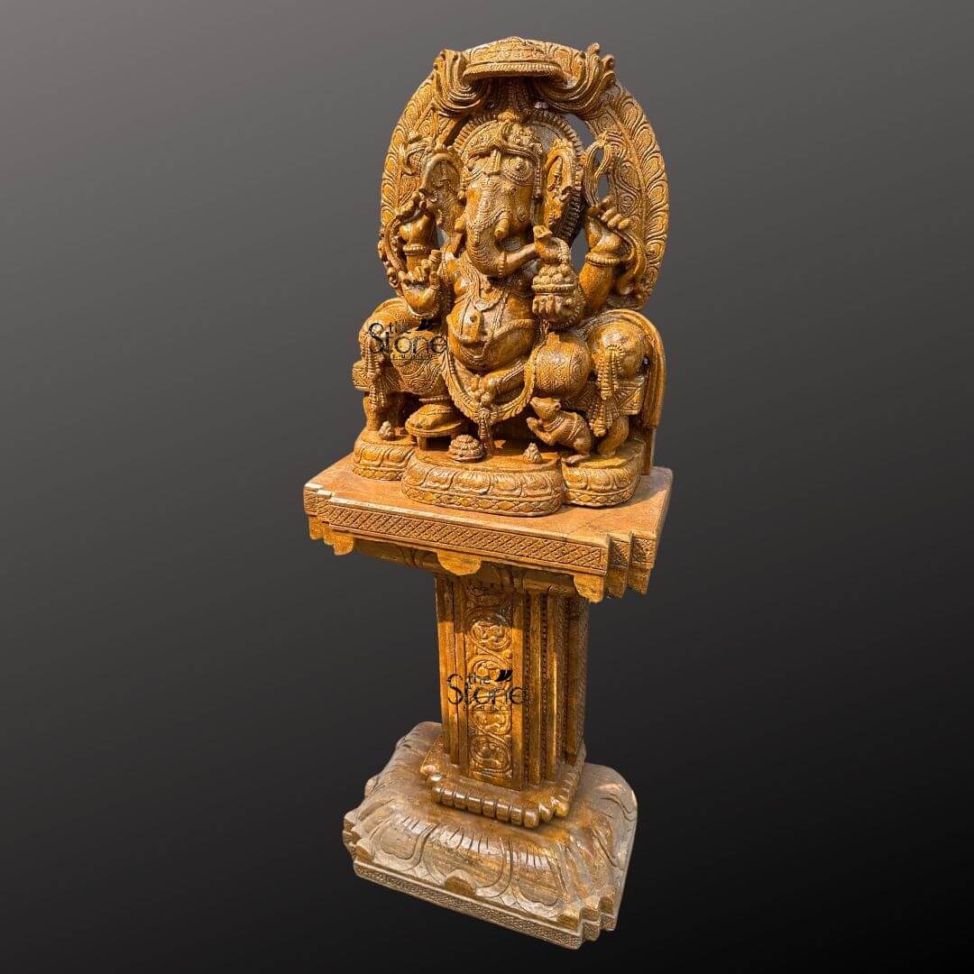 This is a full-view three-quarter angle photograph of a magnificent hand-carved brown sandstone statue of Lord Ganesh elevated and displayed upon a matching ornately carved sandstone pedestal. The statue depicts Ganesh in a traditional seated posture known as Vighneshvara upon a richly carved multi-tiered throne. He is adorned with intricate traditional jewelry a detailed crown and garlands all carved directly from the warm textured brown sandstone. Ganesh has four arms, holding symbolic attributes including an axe and a goad with his primary right hand in the Abhaya mudra of blessing. His form is robust and detailed with a serene facial expression and deeply carved reliefs that showcase artisanal mastery. The Ganesh statue rests upon a substantial square sandstone pedestal base, which is richly carved on all visible sides with traditional Indian motifs including decorative floral patterns miniature figural reliefs and architectural elements that echo the artistry of the main sculpture. The pedestal provides a stable and elegant foundation raising the deity to a prominent height suitable for display in a grand villa entranceway. The entire ensemble—statue throne and pedestal—is crafted from the same consistent brown sandstone, featuring a beautiful uniform natural grain and a finely textured, matte finish that emphasizes the depth and complexity of the hand-carvings. The statue and pedestal are presented against a clean seamless dark grey studio background that fades to black isolating the object and allowing its form color and intricate details to be the sole focus. The lighting is soft even and directional illuminating the scene from the front and slightly to the side to cast gentle shadows that define the three-dimensional quality and deep reliefs of the sandstone carving. The overall composition conveys a sense of spiritual reverence traditional artistry and substantial craftsmanship inherent to this type of premium Indian stone sculpture making it a commanding and welcoming presence for a high-end entrance.