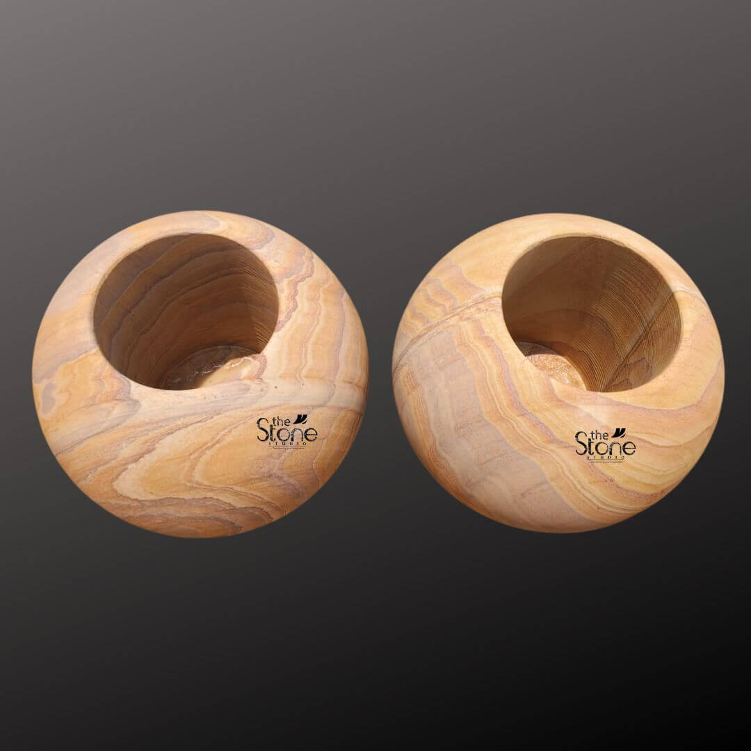 A studio photograph presents a pair of identical spherical sandstone planter pots arranged side-by-side against a softly graduated dark grey background. Each pot is hand-carved from a single block of natural sandstone showcasing a rich warm honey-gold base color interwoven with distinctive flowing layered strata patterns in shades of cream beige and light brown. This unique natural veining gives each piece a one-of-a-kind, artisanal appearance. The pots are perfectly spherical, with a smooth tactile matte finish that emphasizes the inherent beauty of the stone. The top of each sphere is hollowed out to form a perfectly circular deep basin approximately half the diameter of the pot designed to hold a plant and soil. The rim of the opening is clean and smooth leading down into the planting chamber. The texture of the sandstone is clearly visible exhibiting a fine-grained slightly porous quality that is both durable and aesthetically pleasing. The substantial weight and solid form of the pots convey a sense of stability and quality craftsmanship. These round sandstone flower pots are specifically designed to be a sophisticated and organic addition to any home decor. The earthy colors and flowing patterns of the stone complement a wide range of plant life from succulents and cacti to small flowering plants or ferns. The spherical shape is a timeless and elegant design choice that brings a soft natural element to modern or traditional living spaces. The durability of the sandstone makes them suitable for both indoor and outdoor use ensuring they remain a beautiful and functional piece for years to come. Their artisanal quality and natural material highlight a commitment to sustainable and elegant home accents.
