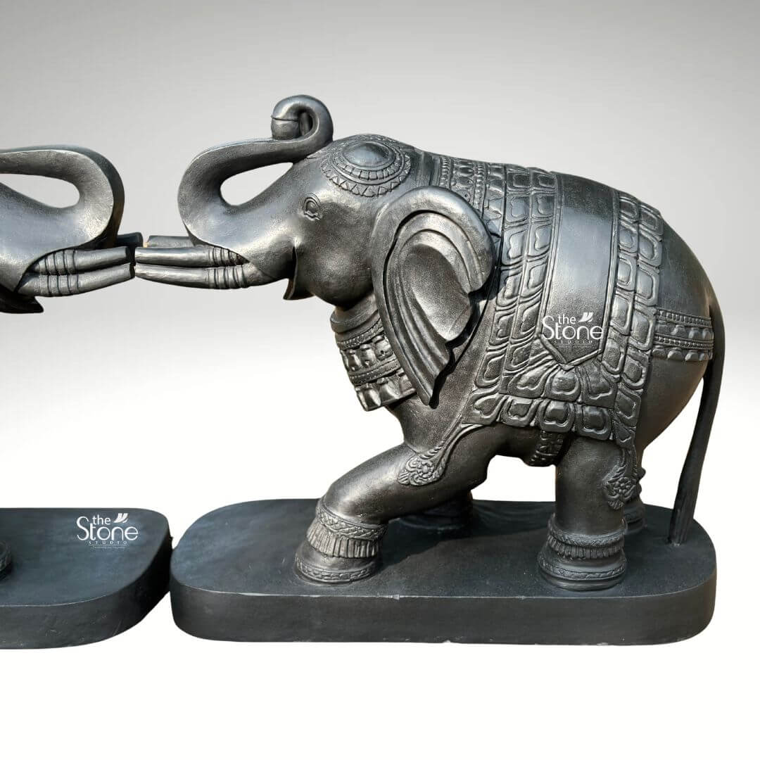 A magnificent pair of 26-inch blackstone elephant statues hand-carved with meticulous care and presented against a neutral light-grey background. The two statues are positioned symmetrically facing each other with their raised trunks touching gently in a traditional gesture of greeting, affection and good fortune. They are sculpted from a dark rich blackstone which has been polished to a smooth matte finish highlighting the deep charcoal color and subtle natural grain of the stone. Each monumental figure is mounted on a simple unadorned black rectangular stone base providing stability and grounding the substantial sculptures. The exceptional artistry of this royal elephant pair statue set is evident in the intricate, fine carvings that adorn their forms. Each elephant is embellished with detailed etched patterns covering their bodies resembling an ornate decorative blanket or howdah cloth complete with traditional Indian motifs scale-like textures and geometric borders. The high level of traditional craftsmanship is further showcased in the meticulous carving of the ears headpieces anklets and the expressive, flowing form of their raised and interlocked trunks. The sharp precise lines of the carvings create a visually rich and tactile surface that catches the light subtly revealing the artisan's skill in rendering lifelike textures from a durable medium. Designed as a commanding and elegant centerpiece for a grand home interior entrywy this blackstone elephant statue pair embodies the grandeur and cultural significance of royal Indian iconography. The dark sophisticated stone material provides a powerful focal point and the symmetrical facing arrangement conveys a deep sene of balance harmony and welcome. This high-quality image effectively communicates the substantial 26-inch scale cultural artistry and spiritual meaning of the sculpture making it an ideal decorative piece for fostering an auspicious and prestigious atmosphere at the threshold of any home.