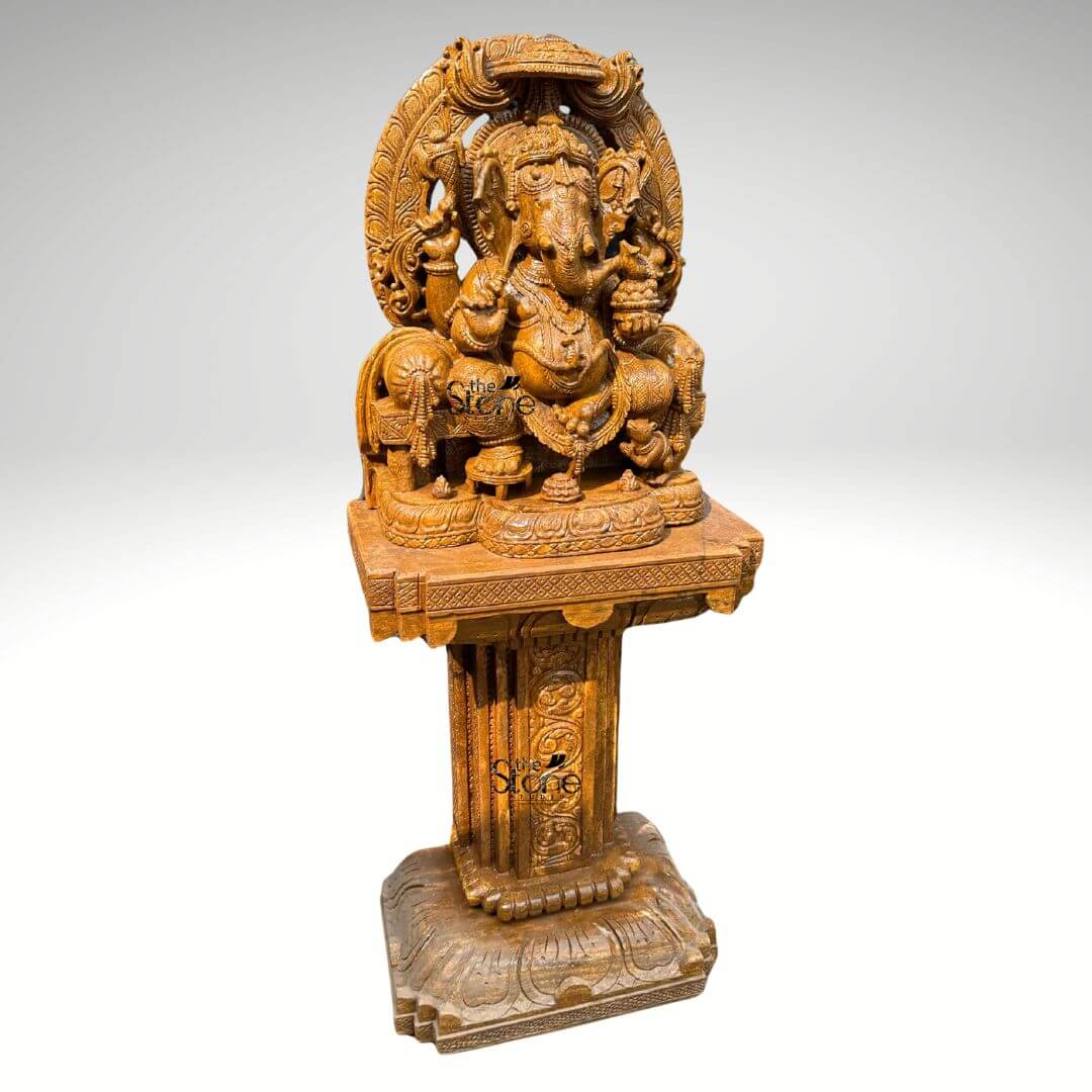 This is a centered three-quarter view photograph of a magnificent, hand-carved brown sandstone statue of Lord Ganesha elevated and displayed upon a matching ornate sandstone pedestal base known as a Peetham. The statue depicts Ganesha in a traditional seated posture known as Vighneshvara upon a richly carved multi-tiered throne. He is adorned with intricate traditional jewelry, a detailed crown and garlands all carved directly from the warm textured brown sandstone. Ganesha has four arms, holding symbolic attributes including an axe and a goad with his primary right hand in the Abhaya mudra of blessing. His form is robust and detailed with a serene facial expression and deep complex carvings that showcase artisanal skill. The Ganesha statue rests upon a substantial square sandstone Peetham base which is richly carved on all visible sides with traditional Indian motifs, including decorative floral patterns miniature figural reliefs and architectural elements that echo the artistry of the main sculpture. The Peetham provides a stable and elegant foundation raising the deity to a prominent height suitable for worship or prominent display as a decor piece. The entire ensemble—statue throne and Peetham—is crafted from the same consistent brown sandstone featuring a beautiful uniform natural grain and a finely textured matte finish that emphasizes the depth and complexity of the hand-carvings. The statue and Peetham are presented against a clean seamless light-grey studio background that fades to white, isolating the object and allowing its form color and intricate details to be the sole focus. The lighting is soft even and directional illuminating the scene from the front and slightly to the side to cast gentle shadows that define the three-dimensional quality and deep reliefs of the sandstone carving. The overall composition conveys a sense of spiritual reverenc traditional artistry and substantial craftsmanship inherent to this type of Indian stone sculpture.