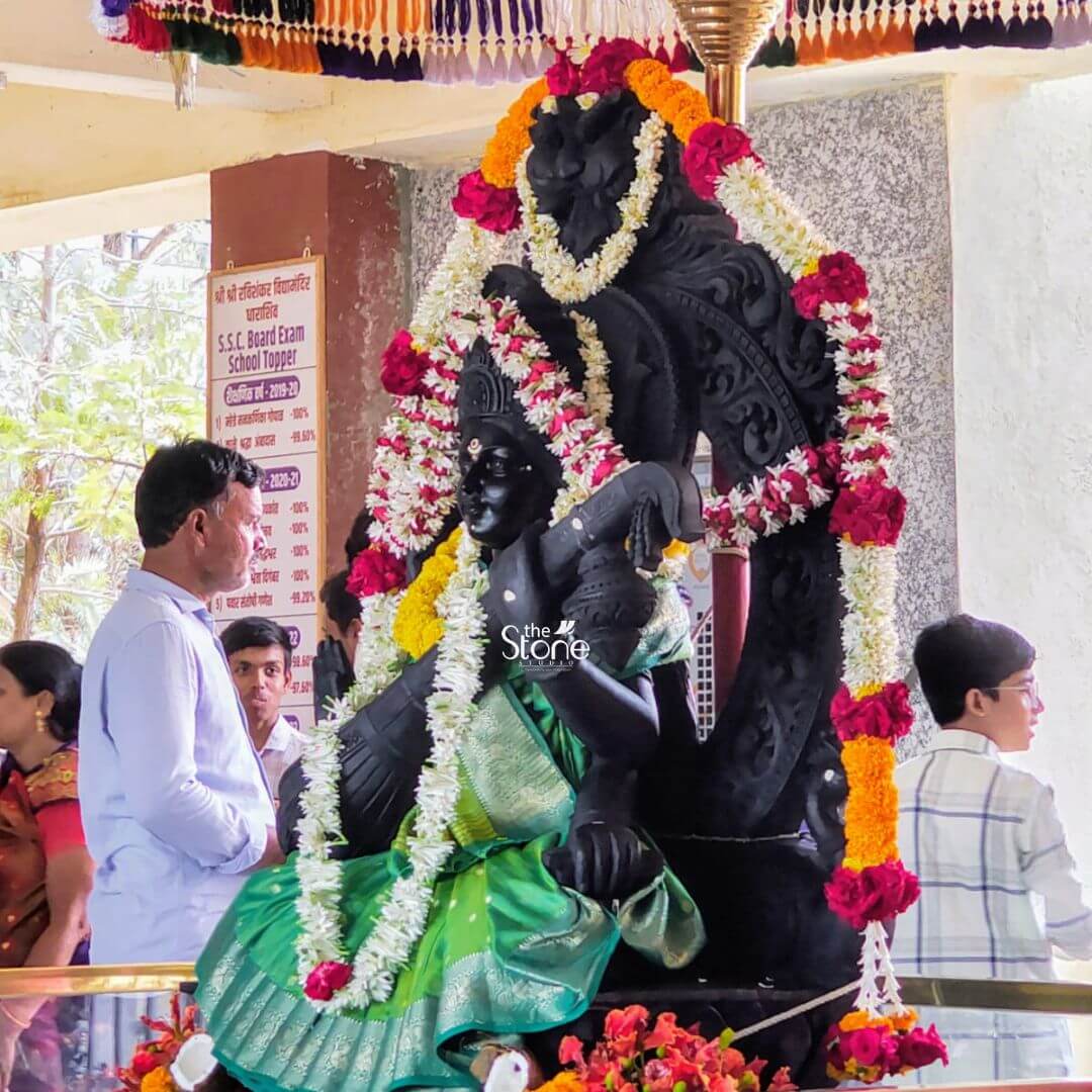 A reverential eye-level photograph captures a magnificent 7-foot Maa Saraswati Saraswati Stone Statue as the divine focal point within a temple setting in Dharashiv Maharashtra. The monumental statue meticulously hand-carved from dark polished blackstone depicts the goddess seated in a graceful traditional posture playing a veena. Her serene face and intricate jewelry are partially visible beneath a profusion of elaborate garlands made of vibrant yellow marigolds and white jasmine which drape heavily from her crown to her lotus base. The statue is enshrined against a decorative carved blackstone backdrop and sits on a flower-covered plinth over which a traditional colorful toran is visible at the top of the frame. The scene is filled with a sense of deep devotion and community worship. In the foreground with their backs to the camera stand two devotees participating in a puja ceremony. To the left a man in a white traditional kurta and topi is seen performing a ritual holding a small brightly burning diya that casts a warm glow on the offerings and the base of the statue. To the right a woman in a blue and gold silk saree stands with her hands joined in pranam facing the goddess. The soft natural light filters in from the open temple mandap illuminating the scene and highlighting the rich textures of the blackstone and the fresh flowers. This image is a profound representation of the Saraswati Stone Statue in Dharashiv showcasing the deep-rooted spiritual and cultural heritage of the region. The sheer scale of the 7-foot idol emphasizes its divine presence towering over the devotees. The meticulous craftsmanship of the blackstone carving combined with the traditional adornments of fresh flowers and silk reflects the artistic and devotional traditions of Maharashtra. The presence of devotees from different generations gathered in worship perfectly illustrates the statue's role as a central point of faith and community tradition making it a fitting visual for an article discussing the significance of this magnificent blackstone murti in Dharashiv.