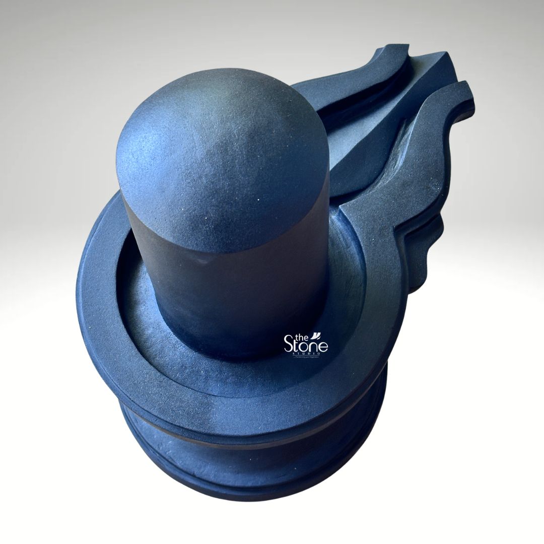 A high-angle studio photograph captures a traditional Blackstone Shivling statue meticulously hand-carved from a single piece of dark rich blackstone. The entire murti rests on a simple rounded pedestal base providing a stable foundation. Rising from this base is the yoni pedestal featuring a smooth perfectly centered shivling shaft. The dark polished surface of the stone absorbs and reflects light subtly emphasizing the heavy, solid presence of the murti and the tactile quality of the material. The clean neutral background ensures that the viewer's attention is entirely on the sculpture's form and texture. The artistry of this Shiva Lingam stone statue is evident in the fine detail and traditional iconography making it ideal for daily Puja. The smooth cylindrical linga represents the formless aspect of Lord Shiva while the yoni base symbolizes the creative energy of Goddess Parvati. The harmonious union of these two elements is central to Hindu philosophy. To the side the intricately carved pranala extends outwards, showing precise channels and traditional decorative motifs designed to catch and channel the ritual offerings of water milk and other liquids during worship. This murti serves as an exemplary representation of a Shiva Lingam Stone Statue for Puja highlighting the specific craftsmanship and quality of the blackstone used. The photograph effectively communicates the cultural significance spiritual symbolism and physical beauty of the devotional object making it a valuable visual reference for devotees interested in acquiring a traditionally crafted Shivling for their home or temple shrine. The high-quality lighting enhances the deep texture of the stone showcasing the skill of the artisan in every curve and line.