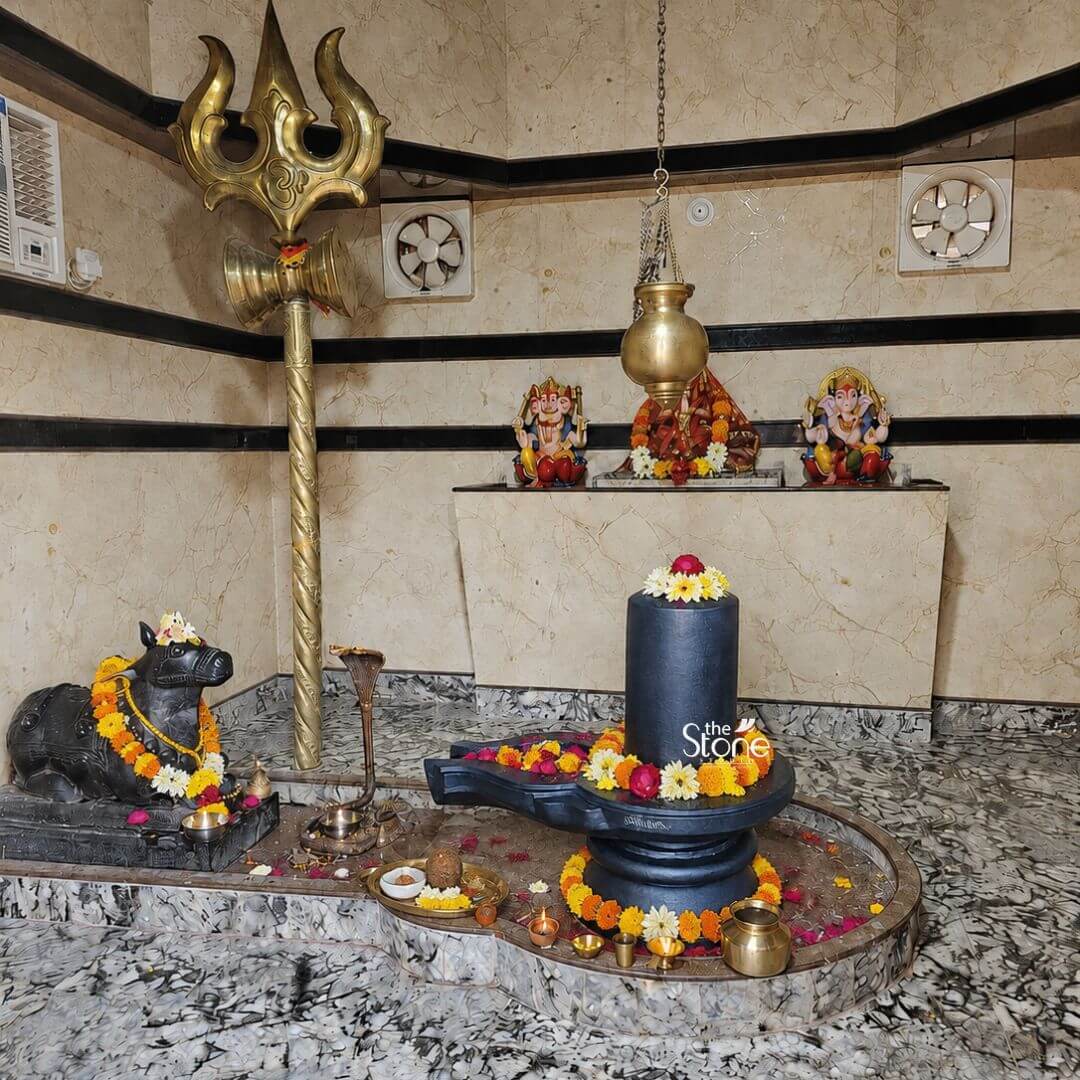 A view inside a home shrine shows a meticulously arranged altar centered around a Blackstone Shivling statue. The murti carved from a single block of dark rich blackstone is the focal point resting on a sculpted yoni base. The cylindrical lingam is adorned with fresh garlands of yellow and orange marigolds and red roses and topped with a single red and yellow flower. The entire Shivling Murti in Black Stone is set within a raised circular marble platform which is also decorated with flower petals brass lamps and a small brass water pot indicating ongoing ritual worship. To the left of the Shivling a black stone Nandi bull statue similarly garlanded with marigolds is seated facing the deity in a pose of reverence. Behind the Nandi stands a tall ornate golden Trishul with a small Damaru attached leaning against the marble-patterned wall. To the right on a raised white marble shelf are three smaller colorful murtis of Ganesha Lakshmi and Saraswati flanked by two circular ventilation fans. A brass ghanti and a panchpatra set are placed on the platform near the Shivling completing the traditional temple setup. This image serves as a perfect illustration for an article about setting up a mandir with a Shivling Murti in Black Stone. It demonstrates the aesthetic and spiritual harmony created by the dark stone murti against the light marble background as well as the traditional placement of associated elements like the Nandi and Trishul. The scene conveys a sense of peace devotion and reverence highlighting the suitability of such a blackstone murti as the central deity in a home place of worship. The rich texture of the stone and the vibrant colors of the floral offerings are clearly visible providing inspiration for devotees.