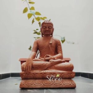 A focused low-angle photograph presents a meticulously hand-carved two-foot-tall Buddha statue made from rich reddish-brown sandstone. The statue is the central element seated in a traditional meditative posture upon an ornate tiered lotus throne which is also carved from the same brown stone. The Buddha is depicted with a serene compassionate expression gently closed eyes and tightly curled hair in the traditional ushnisha. His body is draped in finely detailed monastic robes, the folds of which are sculpted with precision conveying a sense of heavy flowing fabric despite the solid stone medium. His hands are held in the bhumisparsha mudra with the right hand resting on his knee and fingers pointing towards the earth symbolizing his enlightenment. The statue is installed indoors in a minimalist corner placed directly on a dark grey polished stone floor against a clean off-white wall. A single leafy green plant branch possibly a sacred Bodhi tree cutting extends into the frame from the upper left artfully arching behind the Buddha's head and shoulders creating a natural connection and symbolizing growth and wisdom. The background is simple and uncluttered allowing the detailed texture of the porous brown sandstone and the delicate carving of the lotus base to be the main focus. The lighting is soft and natural coming from an unseen source highlighting the sculptural depth and the matte finish of the stone. This image serves as a prime example of a small Buddha stone statue sourced and installed in Bangalore perfectly illustrating the aesthetic appeal and cultural significance of such pieces in a contemporary home environment. The two-foot height makes it an ideal size for a personal shrine living room or meditation space as demonstrated by this setup. The natural brown sandstone with its warm earthy tones complements modern Indian interiors while maintaining a traditional spiritual ambiance. The careful placement combined with the living plant element makes this visual representation highly relevant for articles discussing the selection installation and decoration ideas for Buddha statues in Bangalore residences.