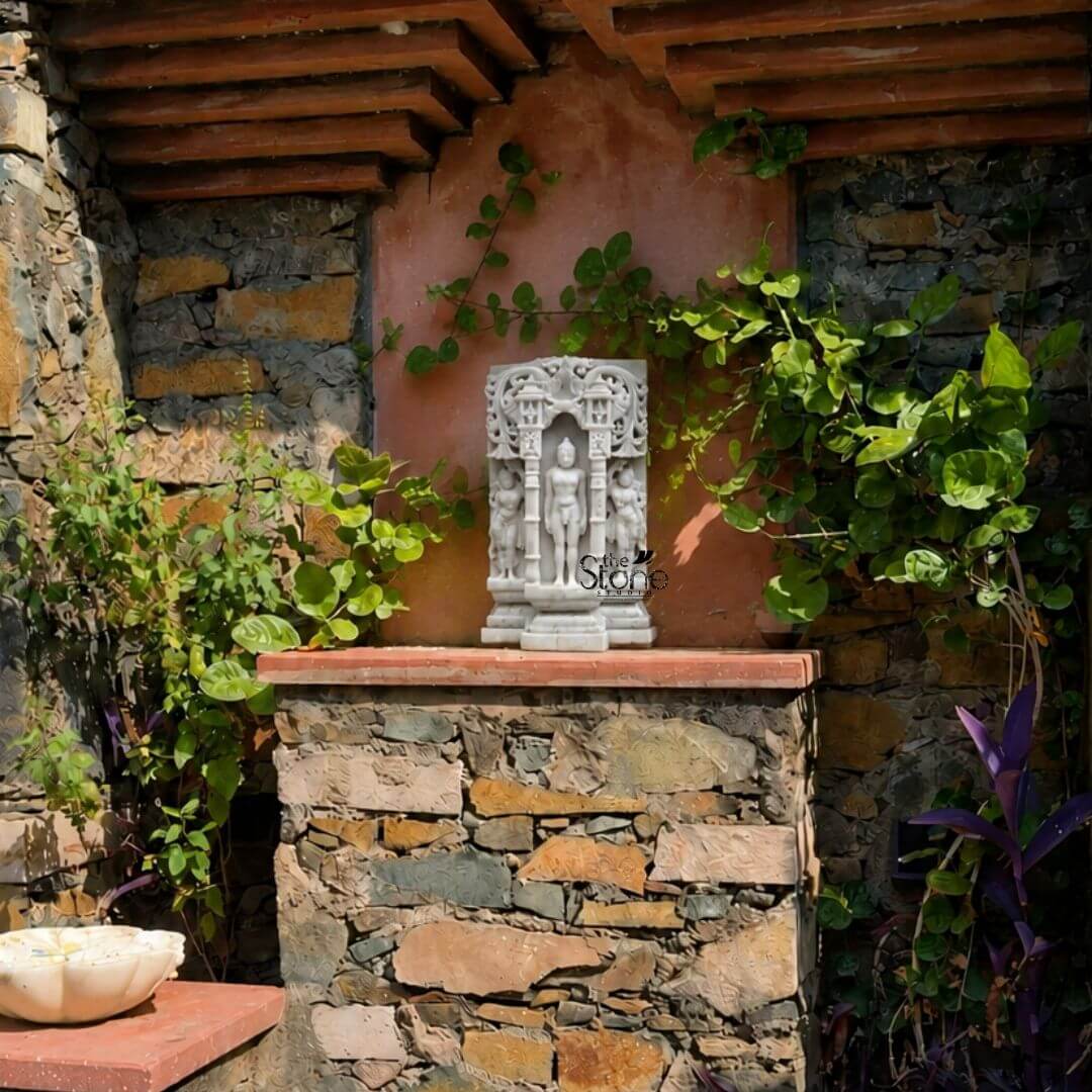 A serene sunlit photograph captures an 18-inch antique-look marble Jain Tirthankara statue standing as a revered centerpiece in a rustic outdoor garden alcove. The statue is meticulously hand-carved from white marble exhibiting a distinct aged patina with subtle color variations and a slightly textured weathered surface giving it the appearance of a centuries-old devotional artifact. The Tirthankara is depicted in the meditative Kayotsarga posture standing perfectly straight and centered within a traditional ornate archway frame or prabhavali which is also carved from the same white marble. The figure is smooth and serene with eyes half-closed in contemplation and elongated earlobes conveying a profound sense of tranquility and detachment. The statue stands on a multi-tiered, stepped marble pedestal which is embellished with carved patterns, further grounding the entire divine composition. The elaborate archway frame surrounding the deity features intricate carvings of traditional motifs including stylized floral scrolls, miniature shrines and two smaller flanking standing figures of yakshas or attendants on either side all rendered with fine detail in the marble. The sculpture's form is balanced and symmetrical exemplifying the high level of craftsmanship involved in creating this devotional artwork. The use of white marble while antiqued symbolizes the purity and clarity central to Jainism. The statue is positioned on a rough-hewn stone plinth against a textured exposed-stone wall which provides a rugged earthy backdrop that contrasts beautifully with the smooth aged marble. The alcove is bathed in natural daylight filtering from the left highlighting the rich textures and sharp lines of the carvings. Lush green climbing vines and various potted plants including a large leaved climber on the right and a smaller leafy plant on the left create a natural sacred grove around the deity. To the lower left a portion of a marble water bowl is visible on a stone shelf.