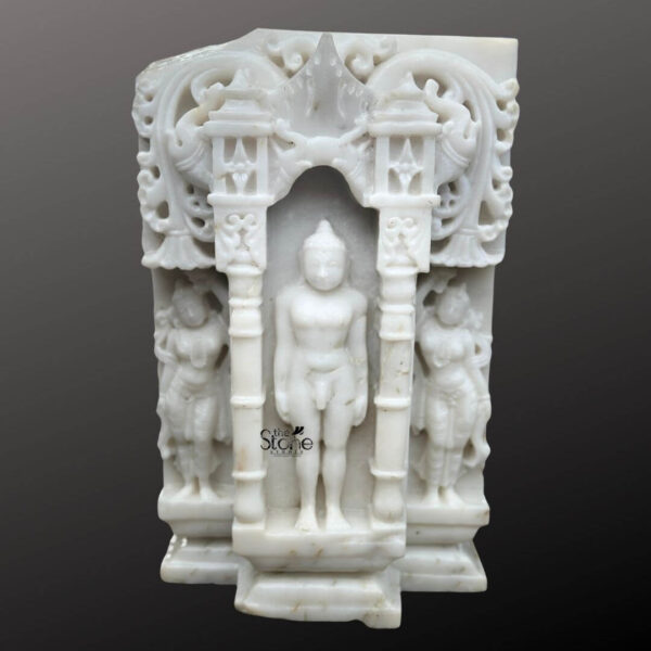 A detailed eye-level photograph captures a magnificent 18-inch Standing Jain Tirthankara Statue meticulously hand-carved from white marble. The statue depicts the Tirthankara in the meditative Kayotsarga posture standing perfectly straight with arms held closely but not touching the body, embodying absolute detachment and tranquility. The figure is centered within a traditional ornate prabhavali frame which is also carved from the same white marble. This elaborate architectural element features a stylized toran adorned with delicate floral motifs miniature shrines and two flanking smaller standing figures of yakshas or attendants on either side of the main deity. The Tirthankara figure itself is smooth and serene with a peaceful facial expression elongated earlobes and the traditional ushnisha at the top of the head. The surface of the white marble exhibits a subtle natural translucency and a fine polished texture highlighting the artisanal skill of the carving. The figure stands on a multi-tiered stepped marble pedestal which is also embellished with carved patterns grounding the entire divine composition. The statue's size makes it a substantial and revered piece suitable for a home altar or a place of worship. This image is an exceptional representation of a Standing Jain Tirthankara Statue in Marble showcasing the rich artistic and spiritual heritage of Jain iconography. The fine details from the delicate carvings on the archway to the serene form of the Tirthankara demonstrate the high level of craftsmanship involved in creating this devotional sculpture. The use of white marble symbolizes purity and clarity core tenets of Jainism making the statue a fitting centerpiece for any article discussing the significance and beauty of such artifacts. The clean dark grey background ensures the white marble figure stands out with clarity allowing every nuance of the carving to be appreciated.