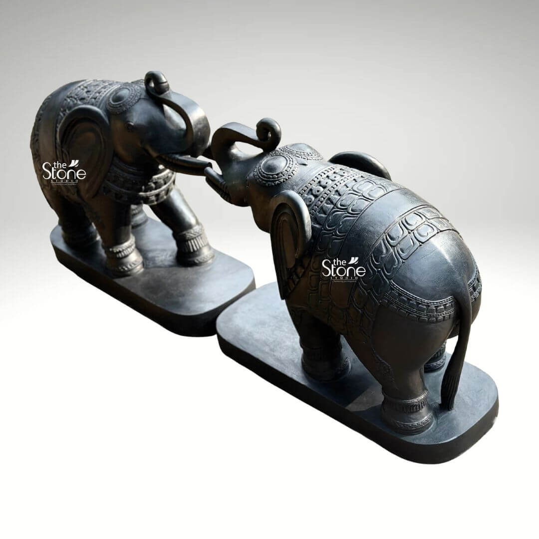 A magnificent pair of 26-inch blackstone elephant statues meticulously hand-carved and positioned symmetrically to face each other. The elephants are sculpted with their raised trunks gently touching, a traditional gesture symbolizing good luck strength and prosperity. They are crafted from a dark rich blackstone giving the entire sculpture a smooth polished texture that highlights the deep charcoal-like color and subtle natural variations of the stone. Both figures are mounted on matching unadorned black rectangular stone bases which ground the monumental figures and provide a stable foundation for display. The artistry of this blackstone elephant statue pair is evident in the fine intricate carvings that adorn their forms. Each elephant is embellished with detailed etched patterns covering their backs resembling an ornate decorative blanket or howdah cloth, as well as on their ears headpieces and legs. The level of traditional Indian craftsmanship is exceptional with sharp clean lines and deep textures that catch the light showcasing the artisan's skill in rendering lifelike forms from a durable medium. From the delicate scale-like patterns on the blankets to the defined wrinkles on the trunks and the gentle curve of the tusks every detail contributes to a sense of grandeur and elegance. Ideally suited as a Stone Elephant Statue Pair for Home & Entrance this 26-inch blackstone sculpture set creates a commanding yet welcoming presence. The dark elegant stone material provides a powerful contrast against a clean neutral light-grey background allowing every nuance of the carving to be appreciated. The symmetrical arrangement and touching trunks convey a deep sense of harmony and balance making this sculpture pair perfect for flanking a grand entryway or serving as a striking focal point in a living space. This image effectively communicates the rich cultural artistry imposing size and spiritual significance of such a decorative piece reinforcing its role in creating an auspicious and spiritually balanced atmosphere within the home.