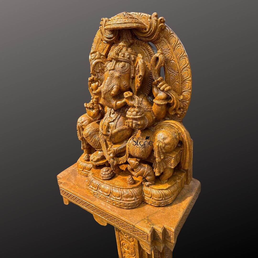 This is a three-quarter view photograph of a magnificent hand-carved brown sandstone statue of Lord Ganesha elevated and displayed upon a matching ornately carved sandstone pedestal designed as a commanding focal point for home decor. The statue is crafted from a warm rich-toned brown sandstone featuring a finely textured matte finish that emphasizes the intricate artisanal craftsmanship. Ganesha is depicted in a traditional seated posture known as Vighneshvara upon a richly carved multi-tiered throne. He is adorned with elaborate traditional jewelry a detailed crown and garlands all carved directly from the stone with deep reliefs. Ganesha has four arms holding symbolic attributes including an axe and a goad with his primary right hand in the Abhaya mudra of blessing. The Ganesha statue rests upon a substantial square sandstone pedestal base which is richly carved on all visible sides with traditional Indian motifs including decorative floral patterns miniature figural reliefs, and architectural elements that echo the artistry of the main sculpture. The pedestal provides a stable and elegant foundation raising the deity to a prominent height suitable for display in a home foyer living room or prayer space. The entire ensemble—statue, throne, and pedestal—is crafted from the same consistent brown sandstone featuring a beautiful uniform natural grain that adds to the sculptural quality and visual appeal. The statue and pedestal are presented against a clean seamless dark grey studio background that fades to black isolating the object and allowing its form color and intricate details to be the sole focus. The lighting is soft even and directional illuminating the scene from the front and slightly to the side to cast gentle shadows that define the three-dimensional quality and deep reliefs of the sandstone carving. The overall composition conveys a sense of spiritual reverence traditional artistry and substantial craftsmanship inherent to this type of large Indian stone sculpture making it a meaningful and aesthetic addition to a home environment.
