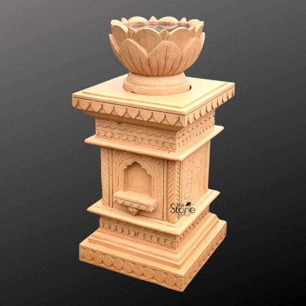 A full-view photograph presents a finely carved sandstone Tulsi Vrindavan planter designed for home use set against a neutral grey gradient background. The Vrindavan is a traditional architectural structure often used for housing the sacred Tulsi plant in Hindu households. This particular planter is crafted from a warm light-peach colored sandstone showcasing a matte textured finish. It stands on a multi-tiered base with the lowest plinth featuring a carved repeating scalloped or petal-like pattern. Above this is a square shaft with a plain central section topped by a small ornate cornice. The main body of the Vrindavan is a square pedestal-like structure adorned with intricate carvings. On the front visible face there is a deeply recessed, arched niche with a small integrated shelf below it. This niche is flanked by vertical panels featuring delicate stylized foliate patterns. The architectural lines are clean yet detailed culminating in a wide overhanging cornice that mirrors the scalloped design of the base. The overall structure conveys a sense of traditional temple architecture on a domestic scale. At the very top resting centrally on the cornice is a separate, deeply carved sandstone element shaped like a blooming lotus flower which serves as the actual planter vessel. The large overlapping petals of the lotus form a bowl-like cavity suitable for holding soil and the sacred Tulsi plant. The entire piece is a cohesive handcrafted item. This Sandstone Tulsi Planter is positioned as a central focal point for a home garden or prayer room emphasizing its role in traditional worship and home decoration. The lighting is even and directional highlighting the depth and detail of the stonework carvings.