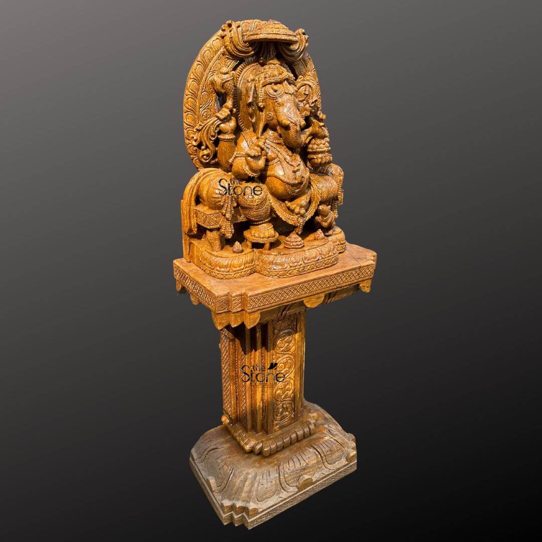 This is a magnificent eye-level photograph of a large hand-carved brown sandstone statue of Lord Ganesha designed in the traditional South Indian temple style and displayed prominently upon an ornate matching pedestal. The Ganesha idol standing approximately five feet tall overall is crafted from a warm rich-toned brown sandstone featuring a finely textured matte finish that emphasizes the deep intricate artisanal carvings. Ganesha is depicted in a seated majestic posture known as Vighneshvara upon a richly carved multi-tiered throne that includes miniature elephant and lion figures. He is adorned with elaborate traditional jewelry a detailed crown and garlands all carved directly from the stone with profound depth and artistry. The Ganesha idol rests securely upon a substantial square sandstone pedestal base which provides a stable and elegant foundation suitable for a temple or grand foyer. This pedestal is richly carved on all visible sides with traditional Indian motifs including decorative floral patterns miniature figural reliefs and architectural elements that echo the art style of the main sculpture raising the deity to a prominent height. The entire ensemble—idol throne and pedestal—is carved from the same consistent brown sandstone showcasing a beautiful uniform natural grain that adds to the sculptural quality and visual appeal. The statue and pedestal are presented against a clean seamless dark grey studio background that fades to black, isolating the object and allowing its form color and intricate details to be the sole focus. The lighting is soft even and directional illuminating the scene from the front and slightly to the side to cast gentle shadows that define the three-dimensional quality and deep reliefs of the sandstone carving. The overall composition conveys a sense of spiritual reverence traditional artistry and substantial craftsmanship inherent to this type of Indian temple-style sculpture.