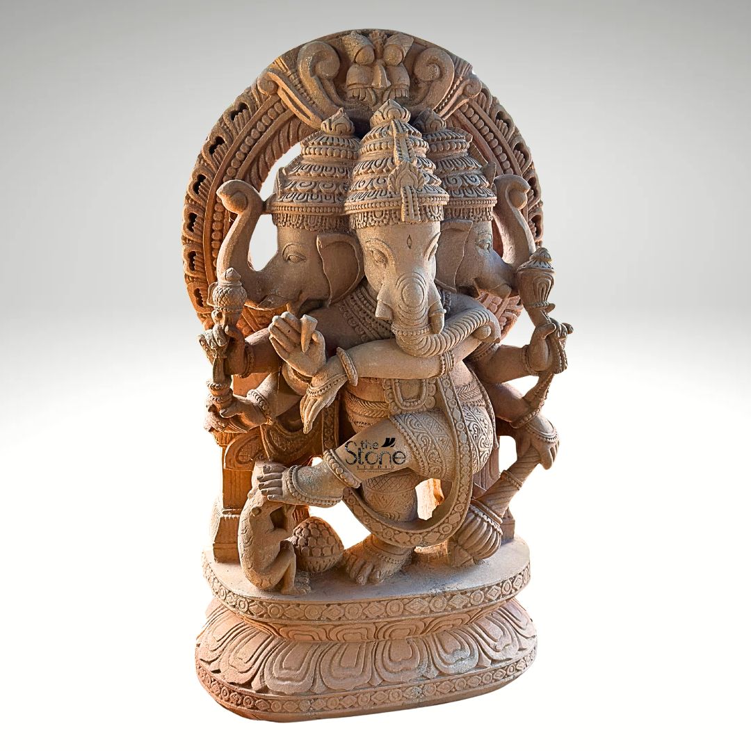 A full-view studio photograph captures a magnificent 4-foot-tall Three Faced Dancing Vinayagar Stone Sculpture hand-carved from a single block of warm, earthy sandstone. The statue is depicted in a dynamic dancing pose with Ganesha's left leg raised and bent gracefully across his body conveying a sense of divine rhythm and movement. He is adorned with elaborate traditional jewelry including multiple necklaces armlets and a sacred thread all intricately sculpted from the same durable stone. The natural porous texture and warm reddish-brown color of the sandstone are beautifully highlighted giving the sculpture a rich tactile quality. This unique murti features three heads with Ganesha's primary elephant face looking forward flanked by two identical elephant faces facing left and right all wearing ornate crowns. Ganesha is shown with eight arms each holding a symbolic attribute such as an ankusha a pasha and a modak with his trunk elegantly curved to taste a sweet from his lower left hand. The sculpture rests on a finely detailed multi-layered lotus throne, grounding the monumental figure. Behind him a large ornate halo with traditional patterns frames his head adding to the divine aura. At Ganesha's feet a small figure of his vahana the mouse is depicted looking upwards in reverence. This image serves as an exemplary representation of traditional Indian stonemasonry and is a perfect illustration for an article discussing the significance of a Three Faced Dancing Vinayagar Stone Sculpture. The photograph highlights the exceptional detail and the imposing presence of the 4-foot sandstone statue making it easy for a devotee or collector to appreciate its spiritual energy and artistic grace. The clean neutral grey background ensures that the viewer's focus remains entirely on the sculpture's form texture and complex iconography conveying the significant role such a murti plays in a home shrine or temple setting.