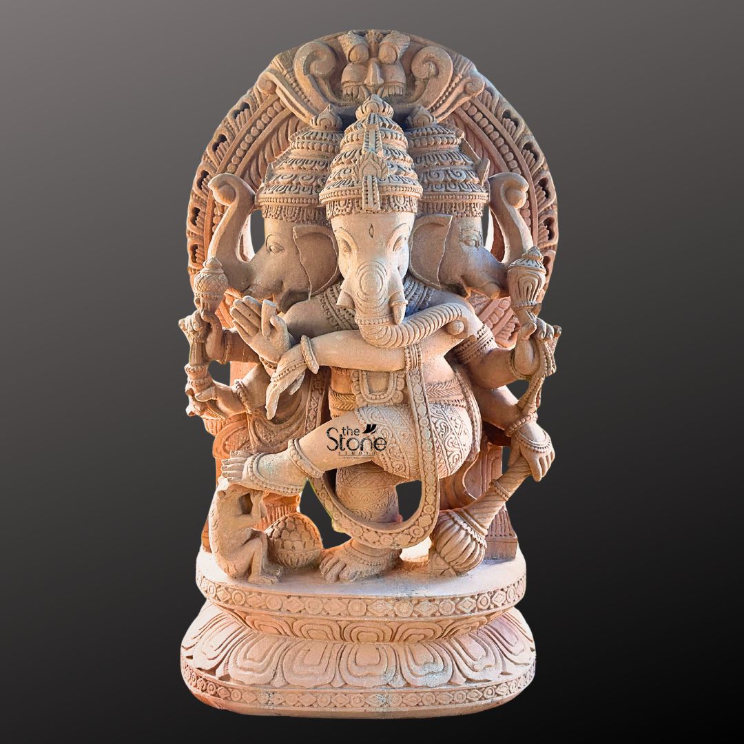 A full-view studio photograph captures a magnificent 4-foot-tall Three Faced Nritya Ganapathi Sculpture hand-carved from a single block of warm earthy sandstone. The statue is depicted in a dynamic dancing pose with Ganesha's left leg raised and bent gracefully across his body conveying a sense of divine rhythm and movement. He is adorned with elaborate traditional jewelry including multiple necklaces armlets and a sacred thread all intricately sculpted from the same durable stone. The natural porous texture and warm reddish-brown color of the sandstone are beautifully highlighted giving the sculpture a rich tactile quality. This unique murti features three heads with Ganesha's primary elephant face looking forward flanked by two identical elephant heads facing left and right all wearing ornate crowns. Ganesha is shown with eight arms each holding a symbolic attribute such as an ankusha a pash and a modak with his trunk elegantly curved to taste a sweet from his lower left hand. The sculpture rests on a finely detailed multi-layered lotus throne, grounding the monumental figure. Behind him a large ornate halo with traditional patterns frames his head adding to the divine aura. At Ganesha's feet a small figure of his vahana the mouse is depicted looking upwards in reverence. This image serves as an exemplary representation of traditional Indian stonemasonry and is a perfect illustration for an article discussing the installation of a Three Faced Nritya Ganapathi Sculpture for Temple. The photograph highlights the exceptional detail and the imposing presence of the 4-foot sandstone statue making it easy for architects temple committees and devotees to appreciate its spiritual energy and artistic grace. The clean neutral grey background ensures that the viewer's focus remains entirely on the sculpture's form texture and complex iconography conveying the significant role such a murti plays in a sacred space.