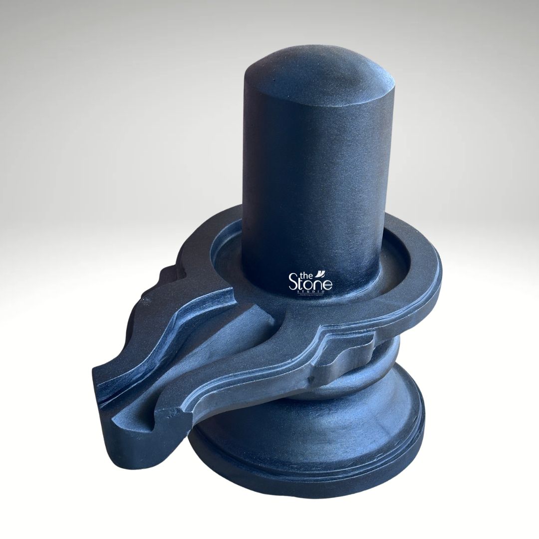 A three-quarter studio photograph captures a traditional Blackstone Shivling statue meticulously hand-carved from a single piece of dark rich blackstone. The murti rests on a simple rounded pedestal base which provides a stable foundation for display. Rising from this base is the cylindrical yoni pedestal featuring a perfectly centered smooth shivling shaft. The dark polished surface of the stone absorbs and reflects light subtly emphasizing the heavy solid presence of the murti and the tactile quality of the material. The clean neutral background ensures that the viewer's attention is entirely on the sculpture's form texture and traditional iconography. The artistry of this traditional black stone Shivling is evident in the fine detail and cultural authenticity. The smooth cylindrical linga represents the formless aspect of Lord Shiva while the yoni base symbolizes the creative energy of Goddess Parvati. The harmonious union of these two elements is central to Hindu philosophy. To the side the intricately carved pranala extends outwards showing precise channels and traditional decorative motifs that are characteristic of fine Indian stonemasonry designed to catch and channel ritual offerings of water milk and other liquids. This image serves as an exemplary representation of a Traditional Black Stone Shivling with Pedestal highlighting the specific craftsmanship and quality of the blackstone used. The photograph effectively communicates the cultural significance spiritual symbolism and physical beauty of the devotional object making it a valuable visual reference for devotees and collectors interested in acquiring a traditionally crafted Shivling for a home or temple shrine. The high-quality lighting enhances the deep texture of the stone showcasing the skill of the artisan in every curve and line.