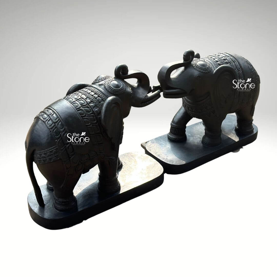 A magnificent pair of 26-inch blackstone elephant statues meticulously hand-carved and positioned facing each other. The elephants are arranged in a traditional affectionate pose with their trunks curled upward and interlocked a gesture often symbolizing greeting strength and good fortune in Indian iconography. They are sculpted from a dark rich blackstone giving the entire sculpture a smooth polished texture that highlights the deep charcoal-like color and subtle natural variations of the stone. Both figures are mounted on matching, unadorned black rectangular stone bases which ground the monumental figures and provide a stable foundation. The artistry of this blackstone elephant statue pair is evident in the fine intricate carvings that adorn their forms. Each elephant is embellished with detailed, etched patterns covering their backs resembling an ornate decorative blanket or howdah cloth complete with traditional Indian motifs scale-like textures and geometric borders. The level of traditional craftsmanship is exceptional with sharp clean lines and deep textures that catch the light showcasing the artisan's skill in rendering lifelike forms from a durable medium. From the delicate patterns on the blankets to the defined wrinkles on the trunks and the gentle curve of the tusks every detail contributes to a sense of grandeur and elegance. Ideally suited for Traditional Elephant Statues for Home Decor this 26-inch blackstone sculpture set creates a commanding yet welcoming presence. The dark, elegant stone material provides a powerful contrast against a clean neutral light-grey background allowing every nuance of the carving to be appreciated. The symmetrical arrangement and touching trunks convey a deep sense of harmony making this sculpture pair perfect for flanking a grand entryway decorating a living space or serving as a striking focal point in a home office. This image effectively communicates the rich cultural artistry imposing size and spiritual significance of such a decorative piece reinforcing its role in creating an auspicious and spiritually balanced atmosphere within the home.