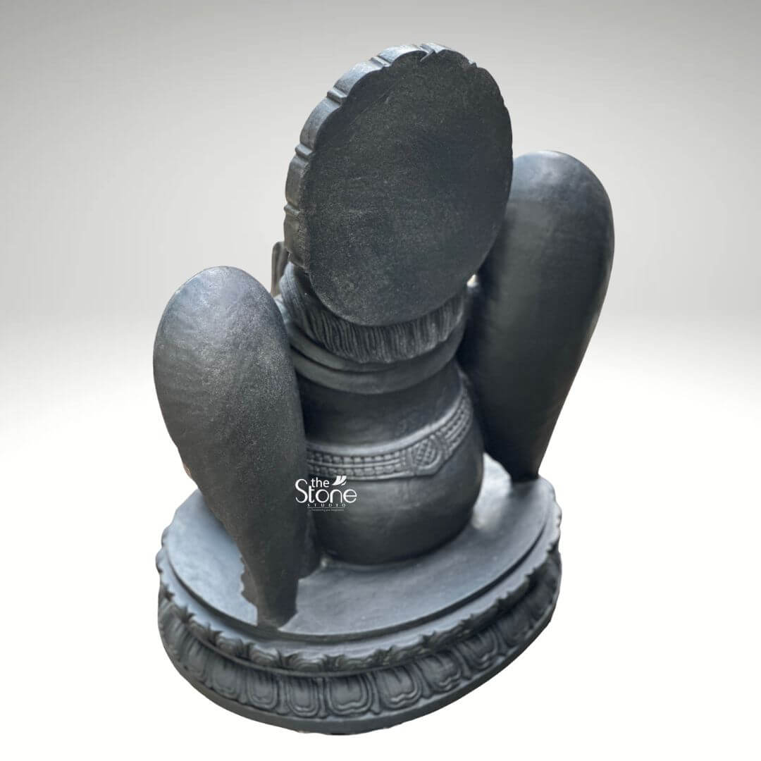 A rear-view eye-level studio photograph presents a meticulously hand-carved black stone statue of Lord Garuda, standing approximately two feet tall against a clean light grey gradient background. The statue is a unified piece of dark polished granite showcasing the stone's natural finely crystalline texture. The view from the back focuses on the intricate sculptural details of the deity's form revealing the elaborate carving of the rear of the traditional crown and the flowing textured hair that cascades down the neck and shoulders all rendered in the dark stone finish. Garuda’s iconic powerful wings are prominent from this angle spread symmetrically behind his torso and exhibiting detailed overlapping feather carving that contrasts with the smooth dark surface of his body. The rear view also clearly displays the sculpted details of his traditional ornaments including the multiple layered necklaces that drape over his shoulders and the intricately patterned waistband or girdle that cinches his waist. The back of his lower garment (dhoti), with its subtle folds and central sash is visible leading down to his kneeling posture on an intricately carved, circular lotus petal base, which provides a stable foundation. This image serves as a vital visual reference for an article discussing the traditional iconography construction quality and spiritual significance of a Traditional Garuda Murti for Temple use. The detailed depiction of the rear craftsmanship particularly the feather work and jewelry patterns is crucial for those seeking authentic, hand-carved devotional art. The two-foot height makes it an ideal size for placement within a temple sanctum or a home shrine representing devotion and protection. The high-quality finish and accurate proportions of this particular statue make it an excellent representation of a traditional Hindu devotional murti.