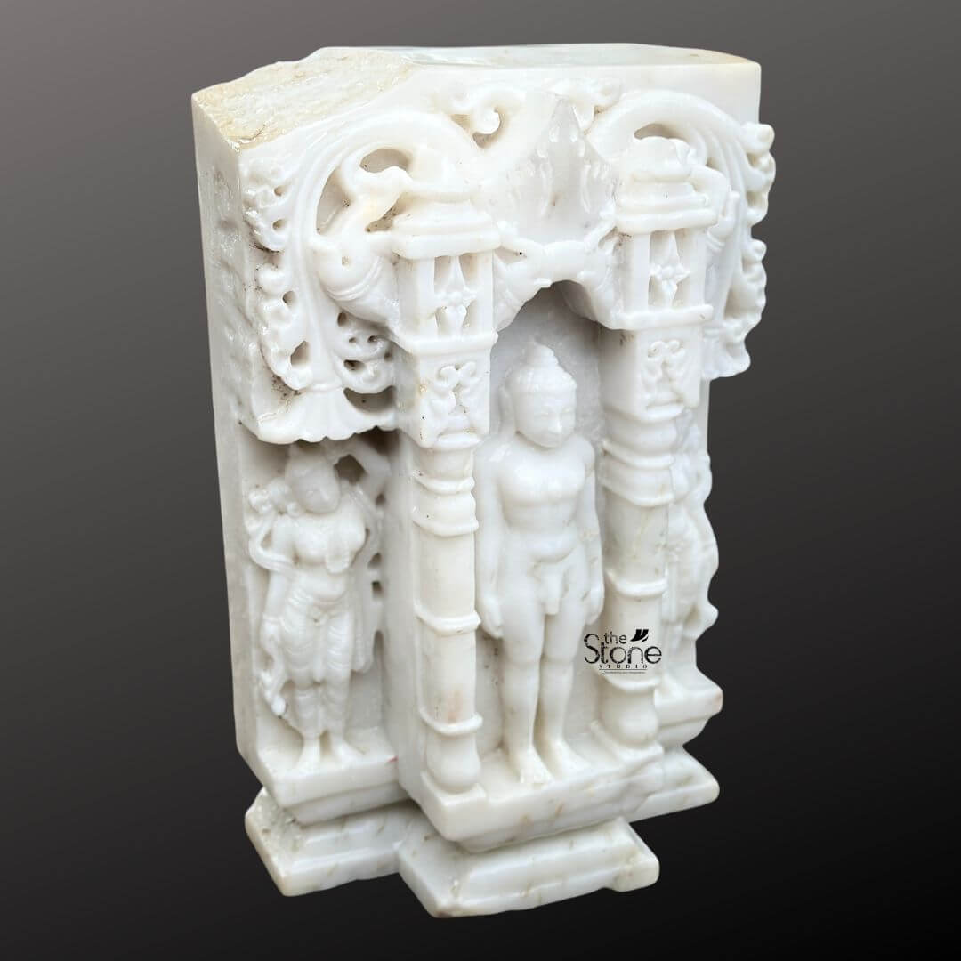 A detailed eye-level photograph captures a magnificent 18-inch Traditional Jain Tirthankara Murti meticulously hand-carved from pristine white marble and featuring a distinct antique-look finish. The central Tirthankara figure is depicted in the meditative Kayotsarga posture standing perfectly straight and centered within a traditional ornate archway frame or prabhavali which is also carved from the same white marble. The figure is smooth and serene with eyes half-closed in contemplation and elongated earlobes conveying a profound sense of tranquility and detachment. The surface of the marble exhibits a subtle aged patina with natural color variations and a slightly textured feel giving it the appearance of a centuries-old devotional artifact consistent with the article's theme of a traditional murti. The Tirthankara stands on a multi-tiered stepped marble pedestal, which is embellished with carved patterns further grounding the entire divine composition. The elaborate archway frame surrounding the deity features intricate carvings of traditional motifs including stylized floral scrolls miniature shrines and two smaller flanking standing figures of yakshas or attendants on either side all rendered with fine detail in the marble. The sculpture's form is balanced and symmetrical exemplifying the high level of craftsmanship involved in creating this devotional artwork. The use of white marble while antiqued symbolizes the purity and clarity central to Jainism. This image is an exceptional representation of a Traditional Jain Tirthankar Murti in White Marble, showcasing the rich artistic and spiritual heritage of Jain iconography. The fine details from the delicate carvings on the archway to the serene form of the Tirthankar demonstrate the artisan's skill in working with this durable and classic medium. The statue's compact 18-inch size makes it suitable for a home altar or a place of worship while its aged aesthetic adds a layer of historical reverence. The clean dark grey background ensures the white marble figure stands out with clarity allowing every nuance of the carving to be appreciated.