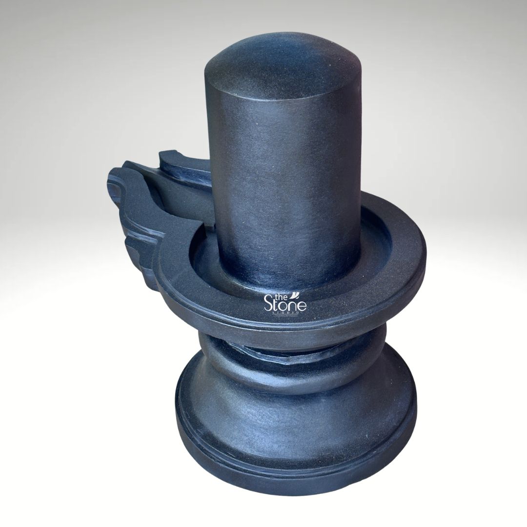 A three-quarter studio photograph captures a traditional Blackstone Shivling statue meticulously hand-carved from a single piece of dark rich blackstone. The entire murti rests on a simple rounded pedestal base providing a stable foundation. Rising from this base is the yoni pedestal featuring a perfectly centere smooth shivling shaft. The dark polished surface of the stone absorbs and reflects light subtly emphasizing the heavy solid presence of the murti and the tactile quality of the material. The clean neutral background ensures that the viewer's attention is entirely on the sculpture's form texture and traditional iconography. The artistry of this traditional Shiva Lingam with yoni base is evident in the fine detail and cultural authenticity. The smooth cylindrical linga represents the formless aspect of Lord Shiva while the yoni base symbolizes the creative energy of Goddess Parvati. The harmonious union of these two elements is central to Hindu philosophy. To the side the intricately carved pranala extends outwards showing precise channels and traditional decorative motifs that are characteristic of fine Indian stonemasonry designed to catch and channel ritual offerings of water milk and other liquids. This image serves as an exemplary representation of a Traditional Shiva Lingam with Yoni Base highlighting the specific craftsmanship and quality of the blackstone used. The photograph effectively communicates the cultural significance spiritual symbolism and physical beauty of the devotional object making it a valuable visual reference for devotees and collectors interested in acquiring a traditionally crafted Shivling for a home or temple shrine. The high-quality lighting enhances the deep texture of the stone showcasing the skill of the artisan in every curve and line.