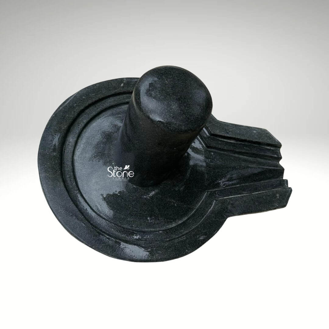 A centered eye-level studio photograph presents a meticulously hand-carved Traditional Shivling Statue made from a solid block of black stone set against a clean light grey gradient background. The entire artifact is a unified piece of dark polished granite showcasing the natural finely crystalline texture and deep rich color of the stone. The central element is the cylindrical Shivling representing the formless aspect of Lord Shiva which rises smoothly and vertically from the center of the circular Yoni pedestal. The top of the Shivling is gently domed and its surface is highly polished creating a subtle sheen that reflects ambient light and contrasts with the matte finish of the surrounding elements. The Yoni base symbolizing the feminine creative energy of Goddess Parvati is a circular basin-like structure surrounding the base of the Shivling. It features a clearly defined raised circular rim and a well-proportioned projecting spout to the right side designed to channel ceremonial offerings of water or milk away from the deity. The entire Yoni base is also meticulously carved from the same dark black stone with precise concentric rings enhancing its form and balance. The carving exhibits traditional craftsmanship with smooth flowing curves and precise edges that give the Shivling and Yoni a harmonious and integrated appearance conveying a sense of ancient sanctity. This image serves as a prime visual reference for an article discussing the traditional iconography and spiritual significance of a Traditional Shivling Statue in Black Stone. The detailed depiction of the dark stone the specific shape of the Yoni and the presence of the spout are all key elements relevant to the article's content. The single-piece construction from durable black granite makes it a sought-after item for daily worship and ritualistic abhishekam in home shrines and temples. The high-quality finish and accurate proportions make this particular statue an excellent representation of a traditional Hindu devotional murti.