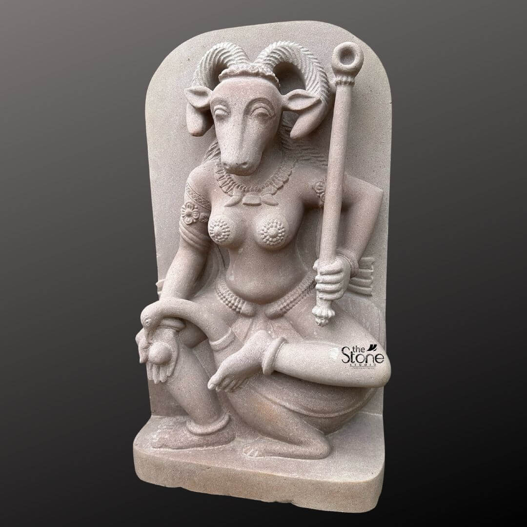 This is a centered full-view photograph of a meticulously hand-carved off-white sandstone statue of the Yogini Vrishanana standing two feet tall and presented against a seamless dark grey studio background that fades to black. The statue is rendered in a high-relief panel style depicting the Yogini a powerful female deity in a complex seated posture with her right leg bent and her foot resting on her mount a recumbent bull. She is sculpted with a human female body and the distinctive head of a horned goat creating the unique Vrishanana form. Her expression is calm and focused gazing forward and she is adorned with deeply carved traditional jewelry including multiple necklaces armlets and a detailed waistband all rendered directly from the stone. The Yogini Vrishanana statue is depicted with four arms showcasing complex iconography. Her primary right hand holds a small fruit or offering while her primary left hand rests gracefully on her raised left knee. In her upper right hand, she holds a water pot and in her upper left hand she firmly grips a ritual staff topped with a symbolic disc. The statue's off-white sandstone material has a finely textured matte finish that emphasizes the high level of detail and the deep reliefs of the carving conveying a sense of age and reverence appropriate for this type of ancient-style art. The statue stands on a simple integrated sandstone base. The composition captures the complete form of the Yogini and her mount presenting the specific iconographic details that define the Vrishanana form. The soft even directional studio lighting illuminates the sculpture from the front and slightly to the side casting gentle shadows that define its three-dimensional form and highlight the rich tactile quality of the sandstone surface. The image accurately represents the two-foot scale of the piece making it a suitable representation for an article discussing this specific traditional Yogini form.