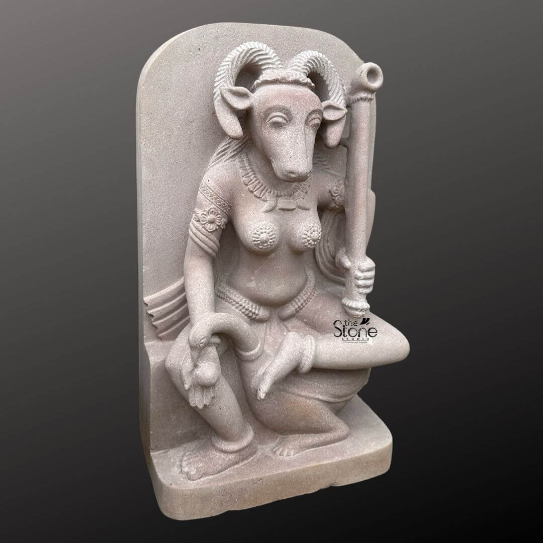 A full-view studio photograph of a meticulously hand-carved off-white sandstone statue of Yogini Vrishanana standing approximately 2 feet tall against a dark neutral gradient background. The statue depicts a female deity with the distinct head of a ram featuring prominent curled ram horns and large attentive eyes. She is seated in a graceful cross-legged Yogic posture on a naturalistic rock plinth with a sculpted fabric drape behind her. Her sculpted body is adorned with traditional Indian jewelry including multiple layered necklaces elaborate armbands with floral motifs wrist bangles and a detailed girdle around her waist. She wears a textured lower garmen. Her upper body is nude with breasts covered by delicate beaded harnesses or covers. In her left hand she firmly holds a ceremonial staff or danda that rests vertically beside her. In her right hand which rests on her bent right knee she holds a small unidentifiable offering or attribute. The entire statue exhibits a soft matte texture typical of hand-carved sandstone with intricate detail visible in the carving of the horns jewelry and facial features. The overall impression is one of ancient spiritual reverence showcasing the traditional iconography of the Yogini cult. The lighting is even and soft highlighting the contours and sculptural depth of the off-white stone. The perspective is eye-level capturing the statue's full presence.