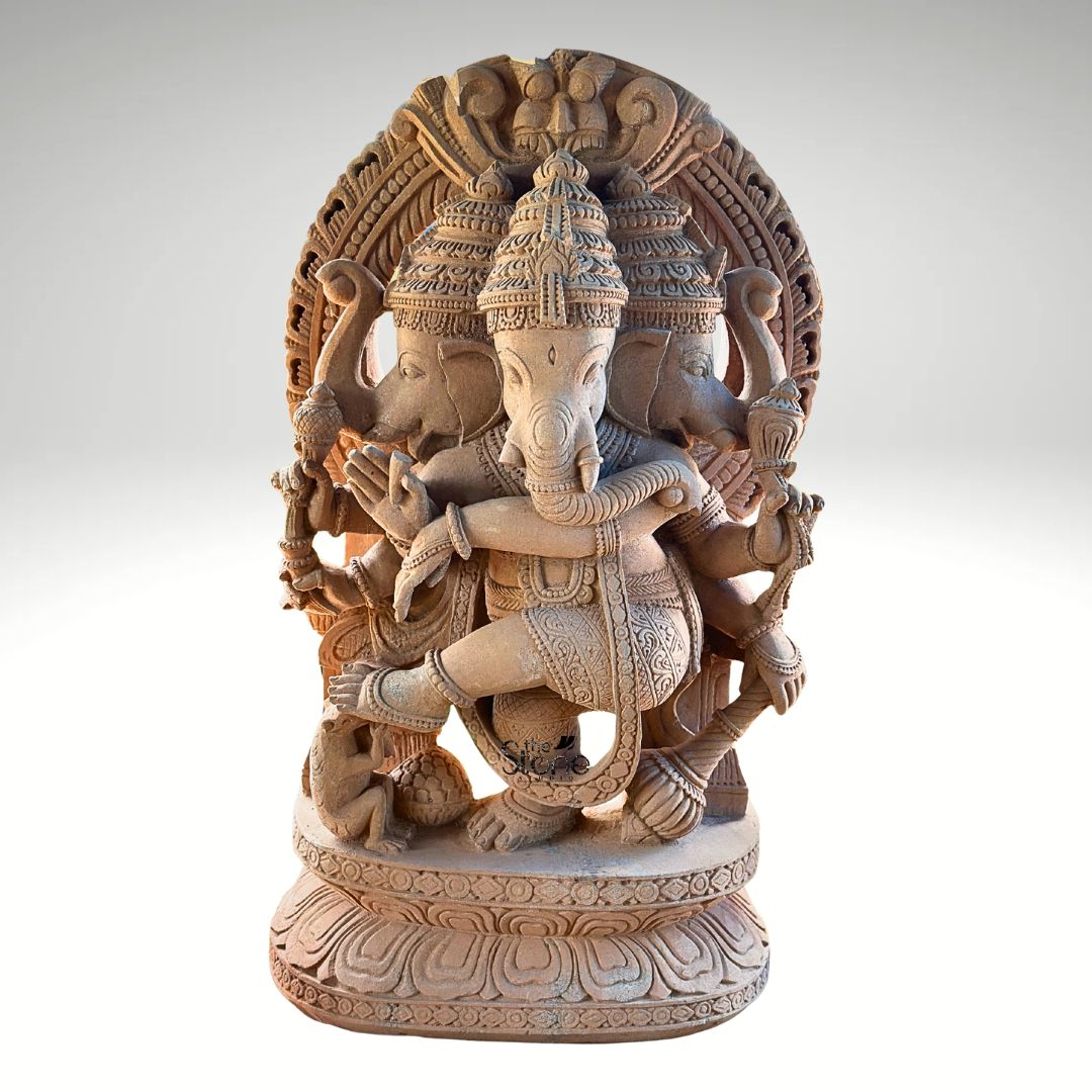 A full-view studio photograph captures a magnificent Trimukha Dancing Ganesha Stone Statue standing approximately 4 feet in height. The statue is meticulously hand-carved from a single block of warm earthy sandstone, giving it a rich porous texture and a solid presence. The central figure of Ganesha is depicted in a dynamic dynamic dancing pose known as Natraja Ganesha with his left leg raised and bent across his body showcasing a sense of movement and divine rhythm. He is adorned with elaborate traditional jewelry including multiple necklaces armlets and a sacred thread all intricately sculpted from the same stone. This unique murti features three heads with Ganesha's primary elephant face looking forward flanked by two identical elephant heads facing left and right all wearing ornate crowns. Ganesha is shown with eight arms each holding a symbolic attribute such as an ankusha and a modak. The main central trunk is elegantly curved to taste a sweet from his lower left hand. A large ornate halo with traditional patterns frames the entire form adding to the divine aura. The statue rests on a finely detailed multi-layered lotus throne grounding the monumental figure. At Ganesha's feet a small figure of his vahana the mouse is depicted looking upwards in reverence. This image serves as an exemplary representation of traditional Indian stonemasonry and is a perfect illustration for an article discussing the significance of a Trimukha Dancing Ganesha Stone Statue. The photograph highlights the exceptional detail and the warm natural color of the sandstone making it easy for a devotee or collector to appreciate the artistry. The clean neutral grey background ensures that the viewer's focus remains entirely on the statue's form texture and complex iconography. This depiction conveys the spiritual energy, cultural artistry, and imposing presence that such a 4-foot sandstone murti brings to a home shrine temple or garden setting.