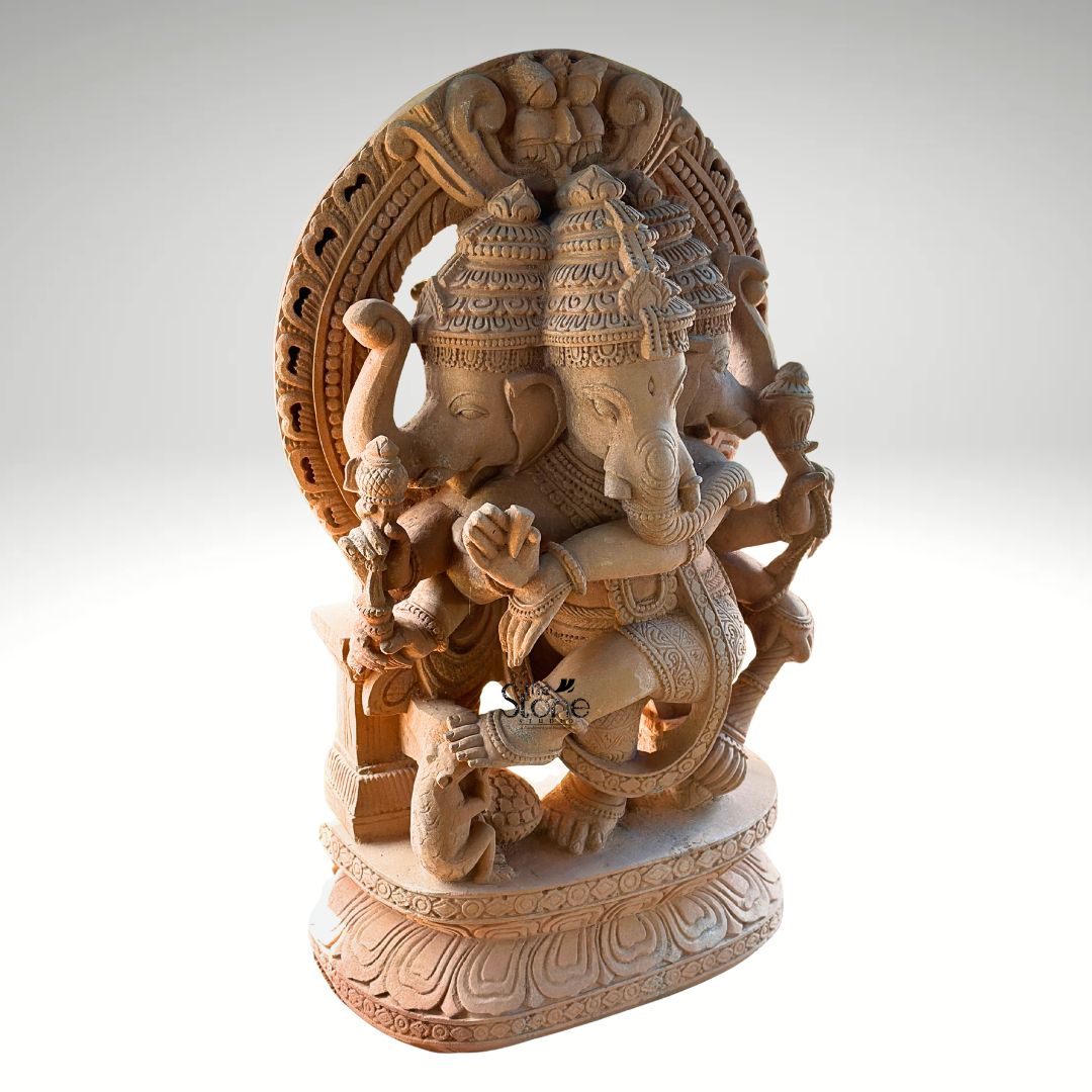 A 4 Feet Ganesha Sandstone statue is shown in a detailed three-quarter view highlighting the sacred form of Trimukhi Vinayagar with three elephant faces carved into one temple-style murti. The handcrafted sandstone sculpture depicts Lord Ganesha seated in a powerful divine posture with multiple arms holding traditional symbolic objects and one hand positioned in a blessing gesture. The warm natural sandstone colour slightly earthy texture and fine hand-carved details give the statue an authentic traditional appearance suitable for temple installation and devotional spaces. The Trimukhi Vinayagar Murti features elaborate crowns on the three Ganesha faces detailed ornaments carved jewellery layered garments and carefully sculpted trunk and facial features. Behind the deity is a large circular prabhavali arch decorated with floral beaded and temple-inspired patterns adding grandeur and sacred presence to the sculpture. The statue is placed on a broad pedestal with lotus-petal carvings and ornamental borders creating a strong foundation that enhances both the spiritual symbolism and visual balance of the 4 Feet Ganesha Sandstone statue. This image connects closely to an article about Trimukhi Vinayagar Murti in Sandstone for Temple Installation by showing the scale craftsmanship, and traditional iconography of the idol. The multi-faced Ganesha form ornate arch detailed arms carved pedestal and natural sandstone finish make the statue ideal for temples mandirs, pooja spaces spiritual centres and sacred architectural settings. The overall design reflects devotion prosperity wisdom and timeless Indian stone carving artistry making this 4 Feet Ganesha Sandstone statue a meaningful choice for temple installation and divine décor.