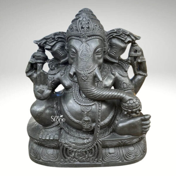 A high-quality studio photograph captures a magnificent Vighnaharta Black Stone Ganesha Statue meticulously hand-carved from a single block of dark rich blackstone. The statue is seated in a traditional posture on an intricately carved pedestal base which is adorned with stylized lotus and geometric patterns. The blackstone has a smooth polished finish that highlights the deep charcoal color and subtle natural variations of the stone giving the murti a rich and heavy appearance. The lighting is even and directional emphasizing the fine details and textures of the carving across the entire form. The craftsmanship of this Blackstone Ganesha statue is exceptional with elaborate detailing evident in every element of the iconography. Ganesha is depicted with his iconic elephant head adorned with an ornate crown elaborate earrings and delicate necklaces that drape over his broad chest. He has four arms his upper right hand holds an ankusha and his upper left hand holds a pasha while his lower right hand is raised in the Abhaya Mudra. His lower left hand gently holds a modak with his trunk elegantly curved to taste it. The large ears are embellished with detailed patterns and his prominent belly is encircled by a decorative udarbandha with a coiled naga acting as the sacred thread. This murti is an exemplary representation of Lord Ganesha as the remover of obstacles embodying both spiritual power and artistic grace. The dark solemn color of the blackstone evokes a sense of reverence and divine presence making it a commanding focal point for a home shrine or temple. The precise carving and traditional iconography make this image a perfect illustration for an article discussing the significance and beauty of such artisanal blackstone Ganesha statues in Hindu tradition. The clean, neutral background ensures that the viewer's attention is entirely on the sculpture's form texture and intricate ornamentation.