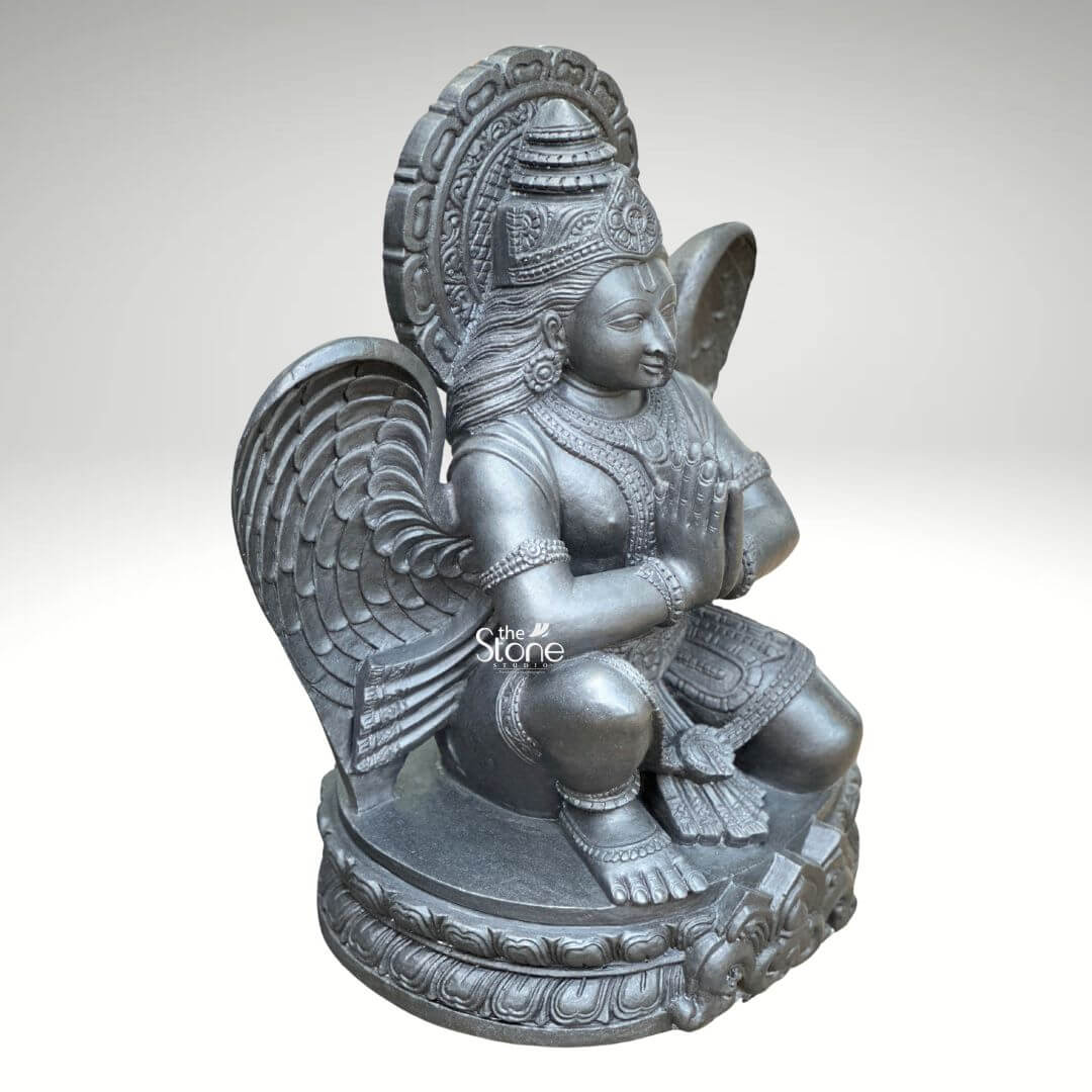 A three-quarter view studio photograph presents a meticulously hand-carved black stone statue of Garud standing approximately two feet tall against a clean light grey gradient background. The statue is a unified piece of dark polished granite showcasing the stone's natural finely crystalline texture. Garuda the divine eagle-man and mount of Lord Vishnu is depicted in a kneeling reverential posture balancing gracefully on his right leg while his left foot rests flat on an intricately carved circular lotus petal base. His hands are pressed together at his chest in the Anjali Mudra conveying deep devotion and respect to his master. His serene human-like face gazes forward with a composed expression framed by elaborate traditional jewelry necklaces armlets wristlets and a tall ornate crown that is further backed by a decorative radiating prabhavali featuring a kirtimukha motif at its apex. Garuda’s iconic powerful wings are fully unfurled and spread symmetrically behind his torso, filling the upper background and exhibiting detailed feather carving that contrasts with the smooth dark surface of his body. He wears a sculpted dhoti with rich patterns and a central flowing sash, all defined by precise lines and intricate detailing that highlight the sculptor's skill. The entire sculpture demonstrates exceptional craftsmanship with sharp clean edges and smooth curves that give the dark granite a fluid lifelike quality as if it were cast in metal rather than carved from stone. The single-piece construction from durable black stone enhances its sense of ancient sanctity and spiritual presence. This image serves as an ideal visual reference for an article discussing the traditional iconography and spiritual significance of a Vishnu Vahana Garuda Statue in Black Stone. The detailed depiction of the dark stone the precise execution of the prayer gesture the elaborate jewelry and the impressive scale of the wings are all key elements relevant to the article's content. The two-foot height makes it a perfect size for home shrines and temple niches representing devotion protection and the divine vehicle of Vishnu. The high-quality finish and accurate proportions make this particular statue an excellent representation of a traditional Hindu devotional murti.