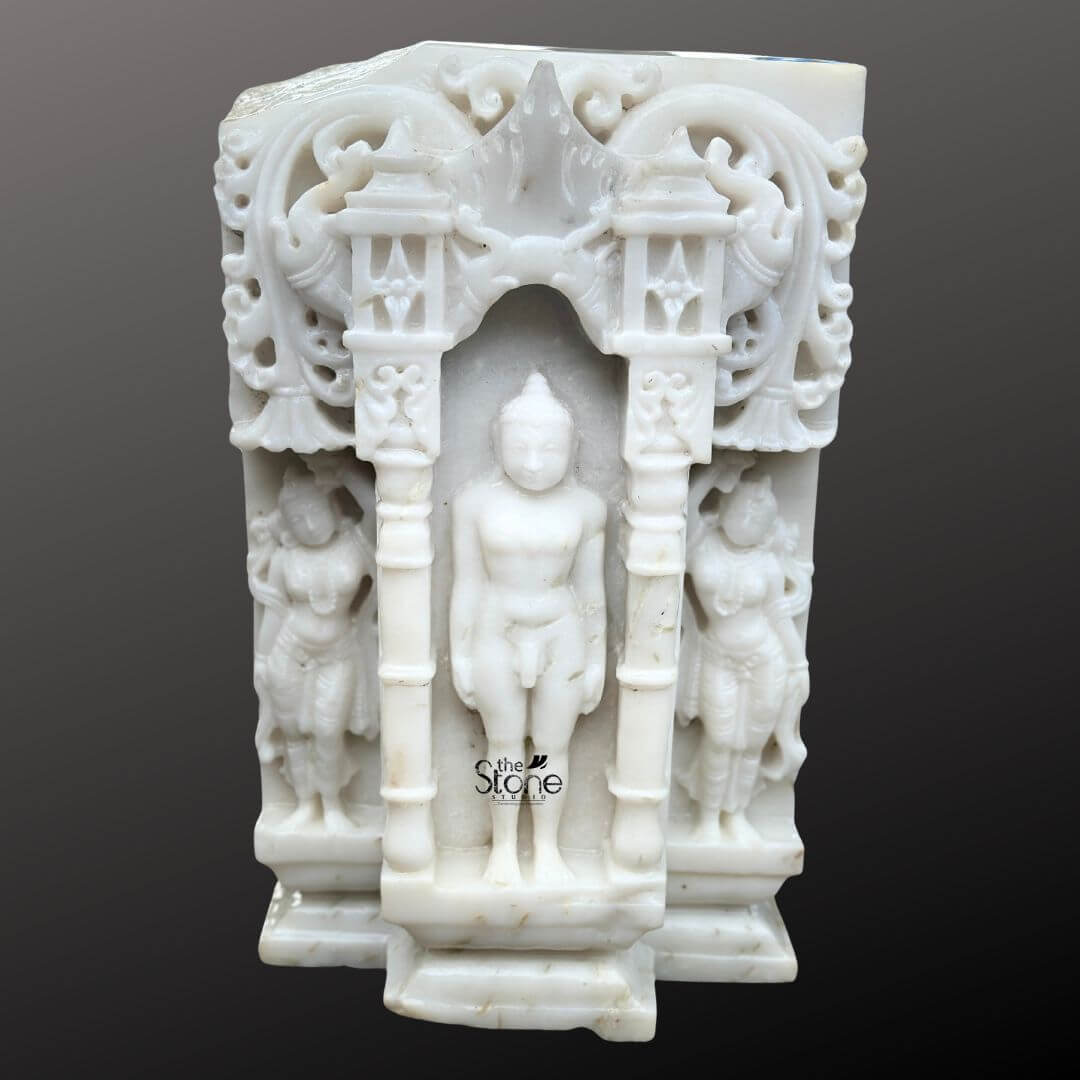 A detailed eye-level photograph captures a magnificent 18-inch White Marble Jain God Idol meticulously hand-carved and featuring a distinct antique-look finish consistent with the article's theme. The central Tirthankara figure is depicted in the meditative Kayotsarga posture standing perfectly straight with arms held closely but not touching the body embodying absolute detachment and tranquility. The figure is smooth and serene with eyes half-closed in contemplation and elongated earlobes. The statue is centered within a traditional, ornate archway frame or prabhavali which is also carved from the same white marble and designed to resemble a miniature temple consistent with the article's theme. The Tirthankara stands on a multi-tiered stepped marble pedestal which is embellished with carved patterns. The elaborate temple-design archway surrounding the deity features intricate carvings of traditional motifs including stylized floral scrolls miniature shrines and two smaller flanking standing figures of yakshas or attendants on either side. The entire composition is balanced and symmetrical exemplifying the high level of craftsmanship involved in creating this devotional artwork. The surface of the white marble exhibits a subtle aged patina with natural color variations and a slightly textured feel giving it the appearance of a centuries-old devotional artifact. This image is an exceptional representation of a White Marble Jain God Idol with Temple Arch showcasing the rich artistic and spiritual heritage of Jain iconography. The fine details from the delicate carvings on the miniature temple archway to the serene form of the Tirthankar demonstrate the artisan's skill. The use of white marble symbolizes purity and clarity core tenets of Jainism making the statue a fitting centerpiece for any article discussing the significance and beauty of such artifacts. The clean dark grey background ensures the white marble figure stands out with clarity allowing every nuance of the carving to be appreciated.