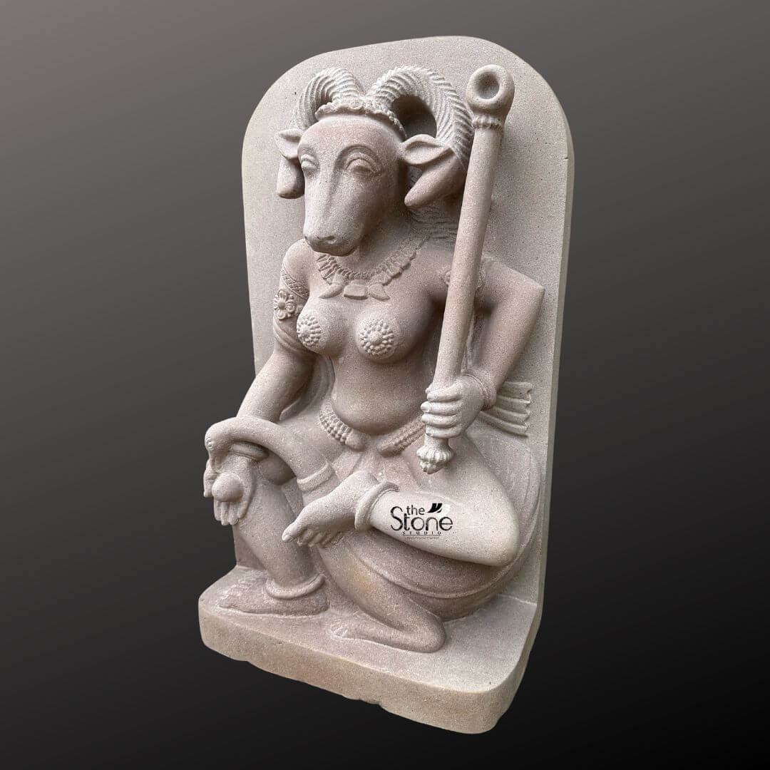 A three-quarter view studio photograph shows a meticulously hand-carved off-white sandstone statue of the Yogini Vrishanan standing approximately two feet tall against a dark neutral gradient background. The statue depicts a female deity with the distinct serene head of a ram featuring prominent curled horns and large calm eyes. She is seated in a graceful cross-legged posture on a rock-like plinth with a sculpted flowing fabric drape serving as a backdrop. Her upper body is adorned with intricate traditional Indian jewelry including multiple layered necklaces ornate armbands with floral motifs wrist bangles and a detailed girdle around her waist. Her breasts are covered by a delicate beaded harness or cover.The Yogini Vrishanana statue holds specific attributes in her four hands although only two are prominently visible from this angle. In her raised left hand she firmly grasps a ceremonial staff or danda that rests vertically beside her. Her right hand resting gracefully on her bent right knee holds a small, indistinct offering or object. The carving exhibits a fine matte texture characteristic of hand-chiseled sandstone with soft even lighting highlighting the sculptural depth and intricate details of her feature the horns and the jewelry. The perspective is eye-level capturing the statue's full presence and delicate carving style. This sculpture represents a fine example of traditional Indian iconography specifically related to the esoteric Yogini cults of medieval times. Vrishanana the ram-faced Yogini is one of the sixty-four Yoginis revered in these traditions. This particular depiction carved from durable off-white sandstone reflects the rich heritage and intricate craftsmanship of Indian temple art. The statue's pose ornaments and ram-headed form are all key elements defining this specific aspect of the divine feminine within the Yogini pantheon. The image serves as a visual reference for articles discussing the historical spiritual and artistic significance of Yogini Vrishanana sandstone statues.