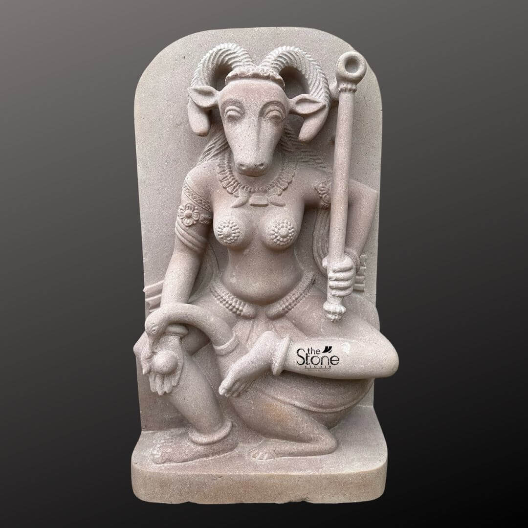 This is a centered full-view photograph of a meticulously hand-carved off-white sandstone statue of the Yogini Vrishanana standing two feet tall and presented against a seamless dark grey studio background that fades to black. The statue depicts the Yogini a powerful female deity in a specific complex seated posture with her right leg bent and foot resting on her mount a recumbent bull. She is sculpted with a human female body and the head of a horned goat creating the unique Vrishanana form. Her expression is calm and focused gazing forward and she is adorned with deeply carved traditional jewelry including multiple necklaces armlets and a detailed waistband all rendered directly from the stone. The Yogini Vrishanana statue is depicted with four arms showcasing complex iconography. Her primary right hand holds a small fruit or offering while her primary left hand rests gracefully on her raised left knee. In her upper right hand she holds a water pot and in her upper left hand, she firmly grips a ritual staff topped with a symbolic disc. The statue's off-white sandstone material has a finely textured matte finish that emphasizes the high level of detail and the deep reliefs of the carving conveying a sense of age and reverence appropriate for this type of ancient-style art. The statue stands on a simple integrated sandstone base. The composition captures the complete form of the Yogini and her mount presenting the specific iconographic details that define the Vrishanana form. The soft even directional studio lighting illuminates the sculpture from the front and slightly to the side casting gentle shadows that define its three-dimensional form and highlight the rich tactile quality of the sandstone surface. The image accurately represents the two-foot scale of the piece making it a suitable representation for an article discussing this specific Yogini form.