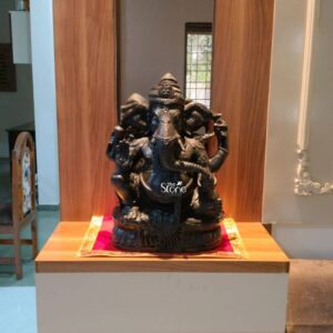 A medium shot photograph capturing an intricately carved 18" Black Stone Ganesh Statue standing centrally on a warm light wood shelf. The statue hand-carved from a rich deep black stone depicts the deity in a traditional seated posture with one leg folded and the other pendant. Its trunk curves gracefully to the left holding a modak. The natural texture of the black stone is visible showcasing the artisan's detailed work on the deity's elaborate ornaments crown and four arms which hold symbolic attributes like an axe and a lotus. The statue rests directly on a simple integrated plinth made of the same black stone. The Ganesh statue is adorned for worship with a fresh garland of small bright pink and white artificial flowers that drape around its neck and chest contrasting beautifully with the dark stone. A small delicate white flower adorns the top of its ornate crown. The statue is centered on the wooden shelf which has a warm grain and is set against a background featuring vertical wooden panels on either side creating a sense of a dedicated shrine space. Below the statue a small red fabric mat with gold embroidery is spread holding the plinth. The background behind the shrine area is softly blurred revealing a modern interior living space typical of a home in Pune. The wooden panels frame the deity making it the clear focal point. The lighting is warm and focused highlighting the deep black color and the fine details of the carving. The overall composition conveys a sense of modern spirituality and reverence making it an ideal centerpiece for home decor. The careful integration of the statue with the natural wood and the traditional textile reflects a common and beautiful home shrine setup in Pune.